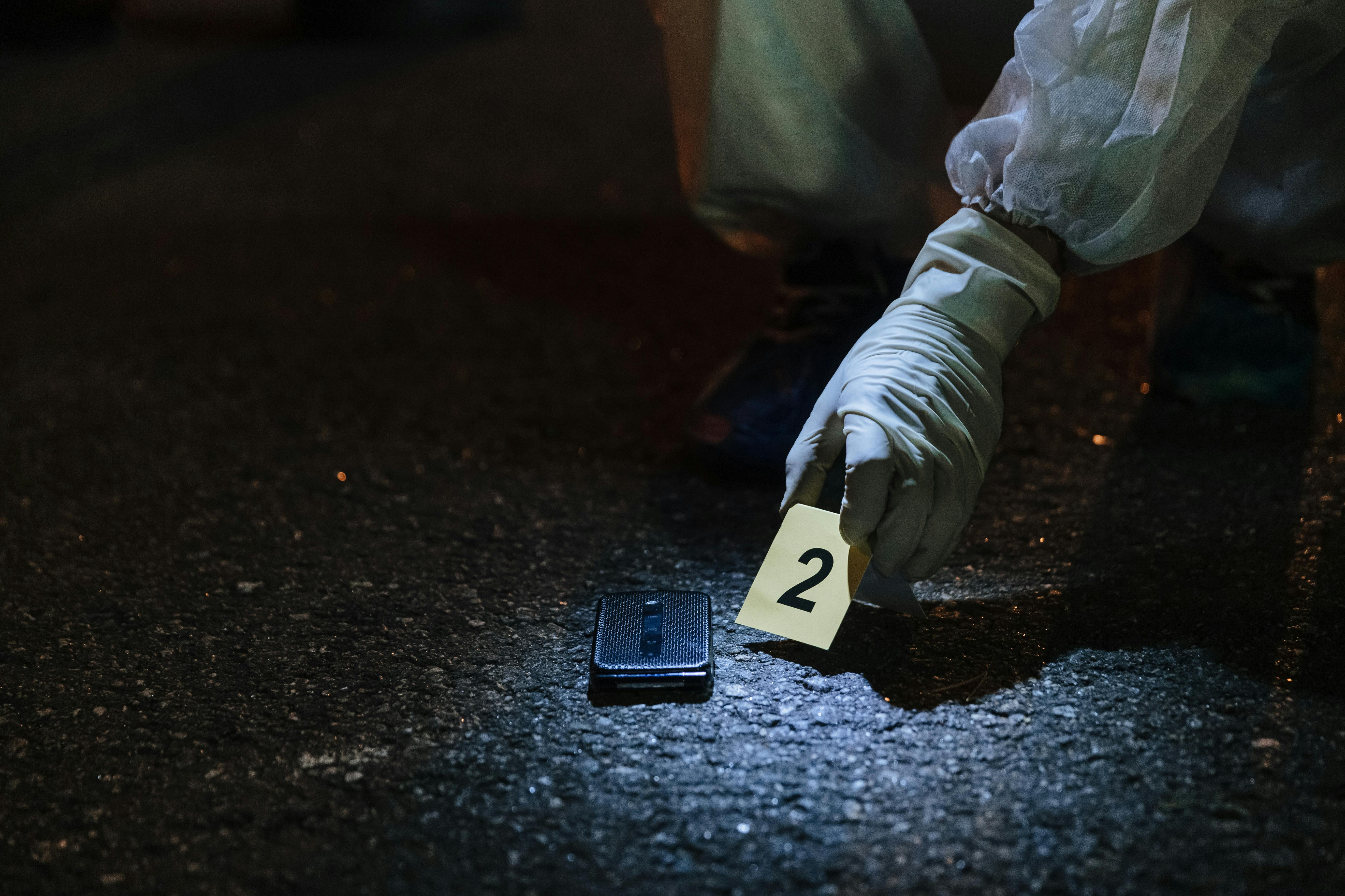 A forensic examiner wearing white gloves and protective gear places a numbered evidence marker next to a dark shoe sole on a wet asphalt surface during evidence collection.