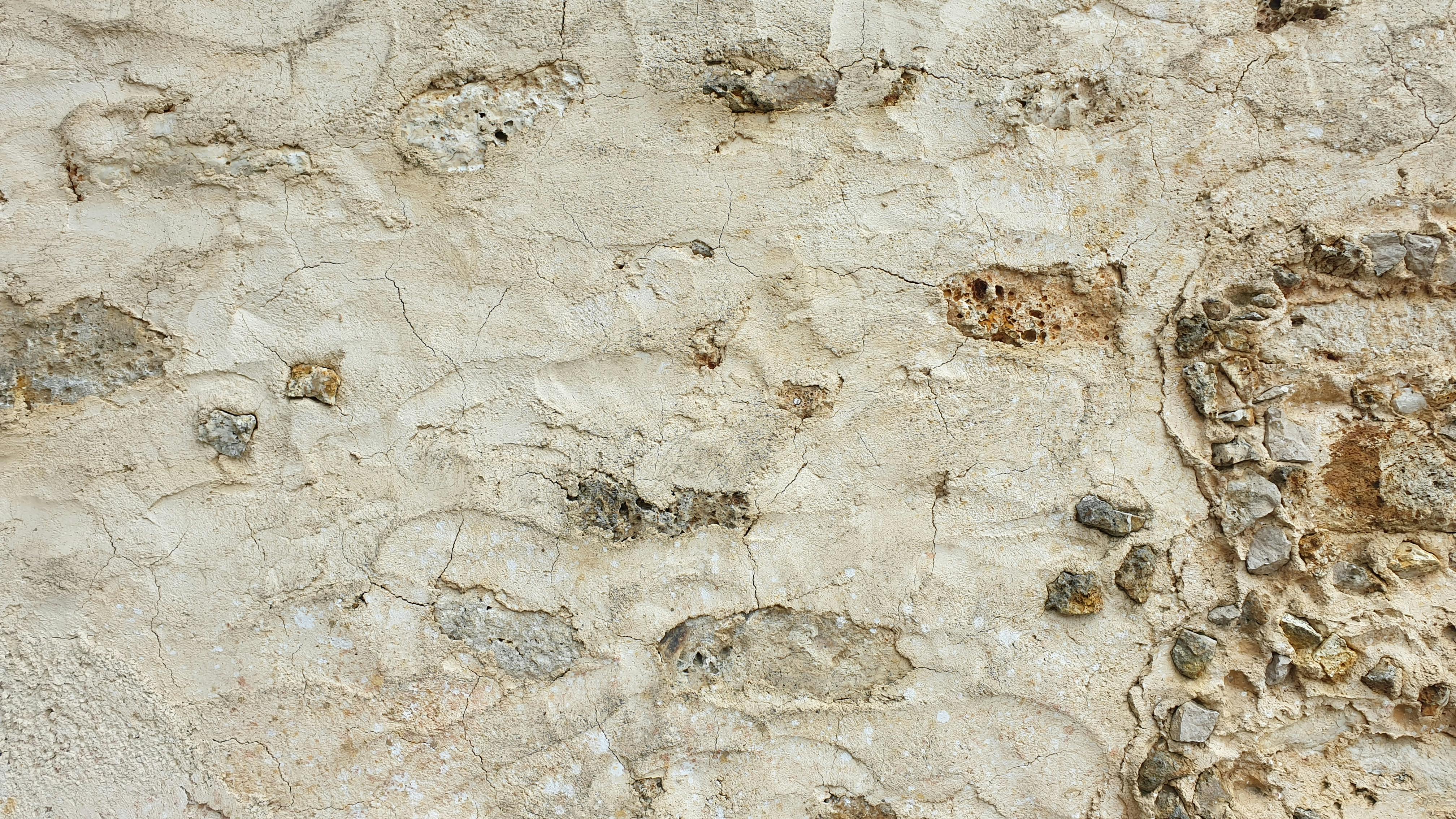 Close-up photograph of a textured stone or aggregate wall surface showing natural material composition with varying earth tones and embedded pebbles or gravel in a light beige and tan palette.
