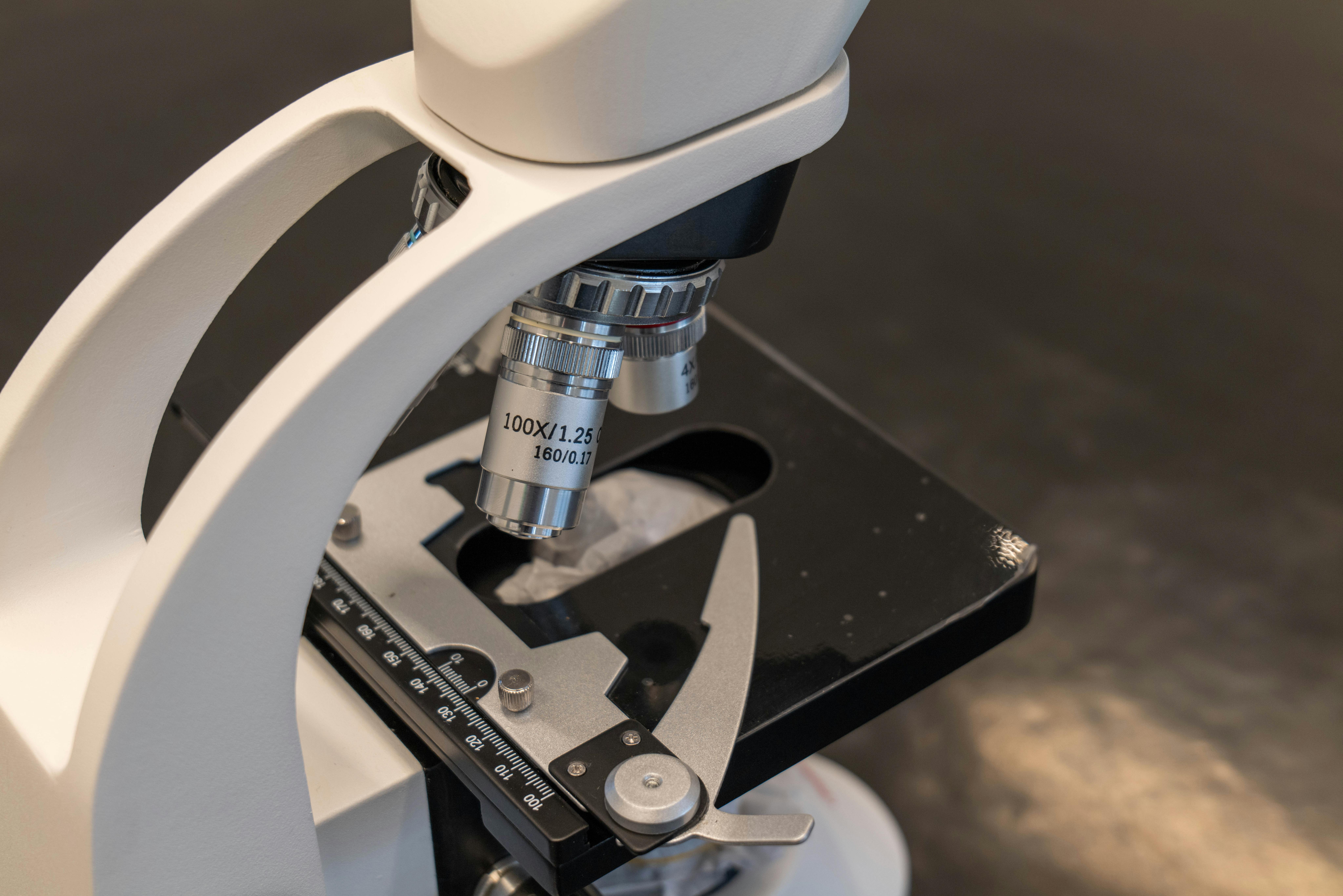 A white compound microscope with a black stage and objective lens positioned over a black specimen platform, photographed from an angled perspective against a neutral background.