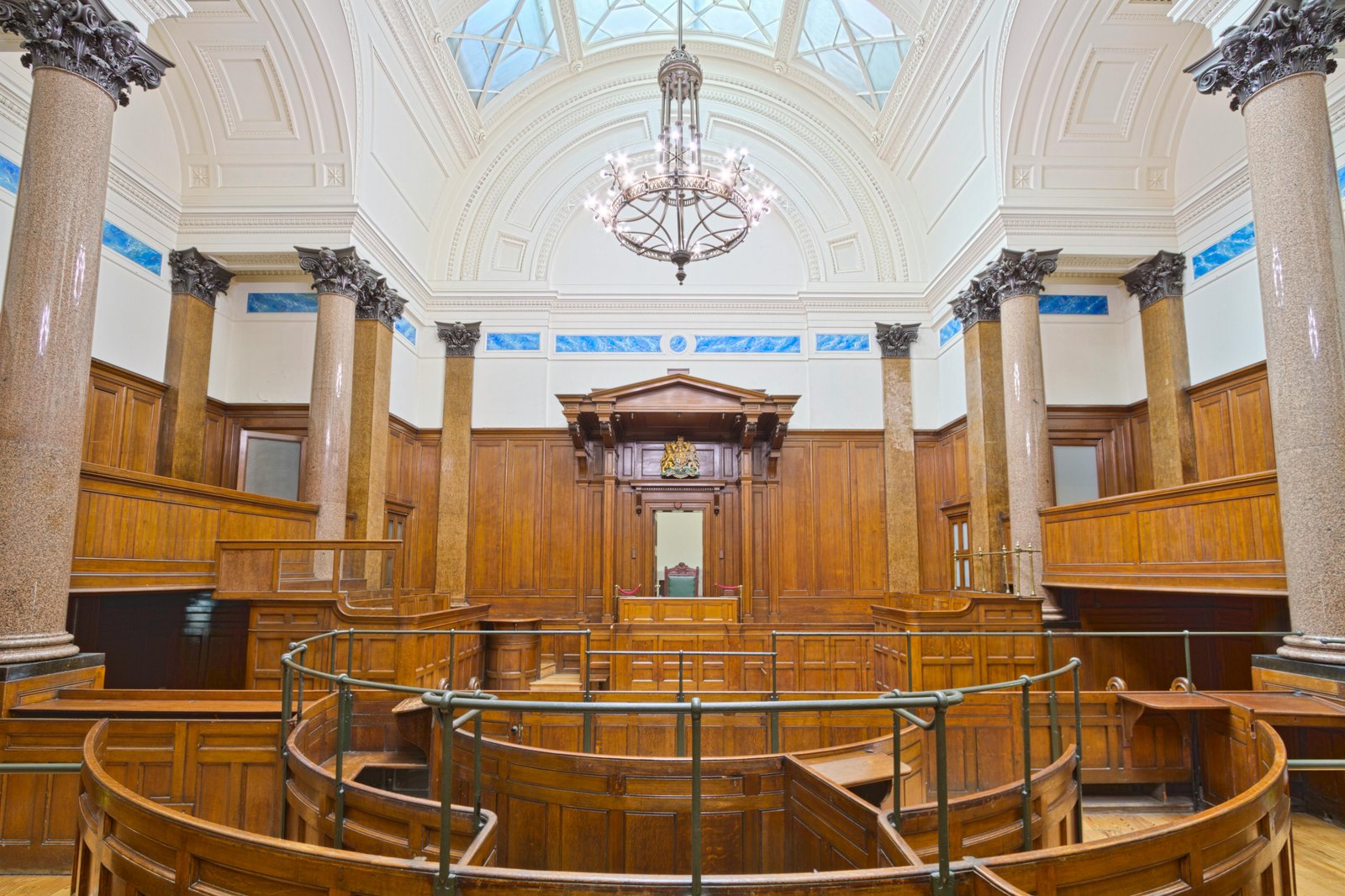 A formal courtroom interior featuring wooden paneling, ornate columns, a vaulted ceiling with skylight, a central judge's bench, and curved jury seating arranged in a semicircular layout.