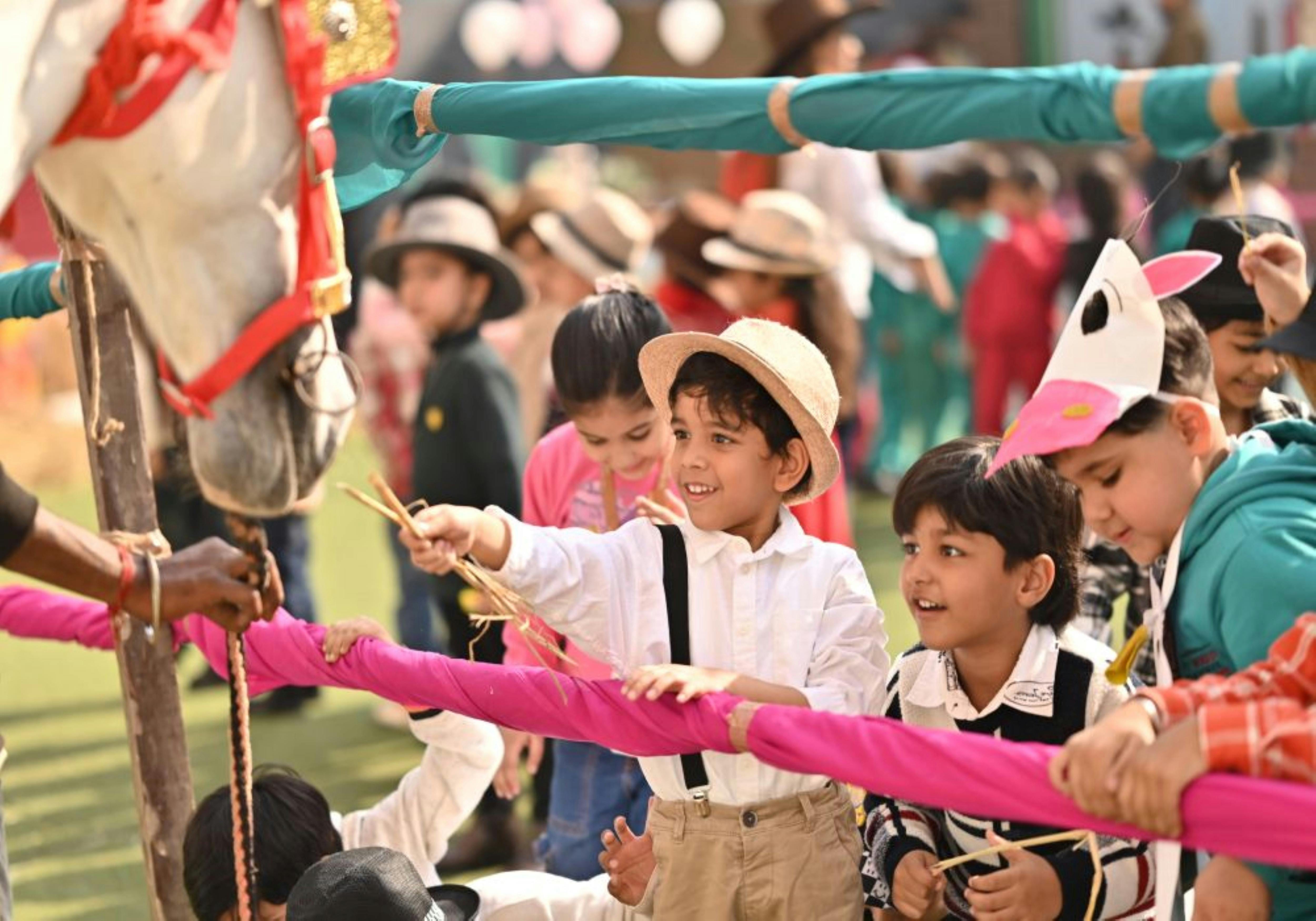 A group of young children wearing hats and casual clothing hold a long pink rope together outdoors, smiling and engaged in a collaborative group activity under a teal-striped canopy with other children and adults visible in the background.