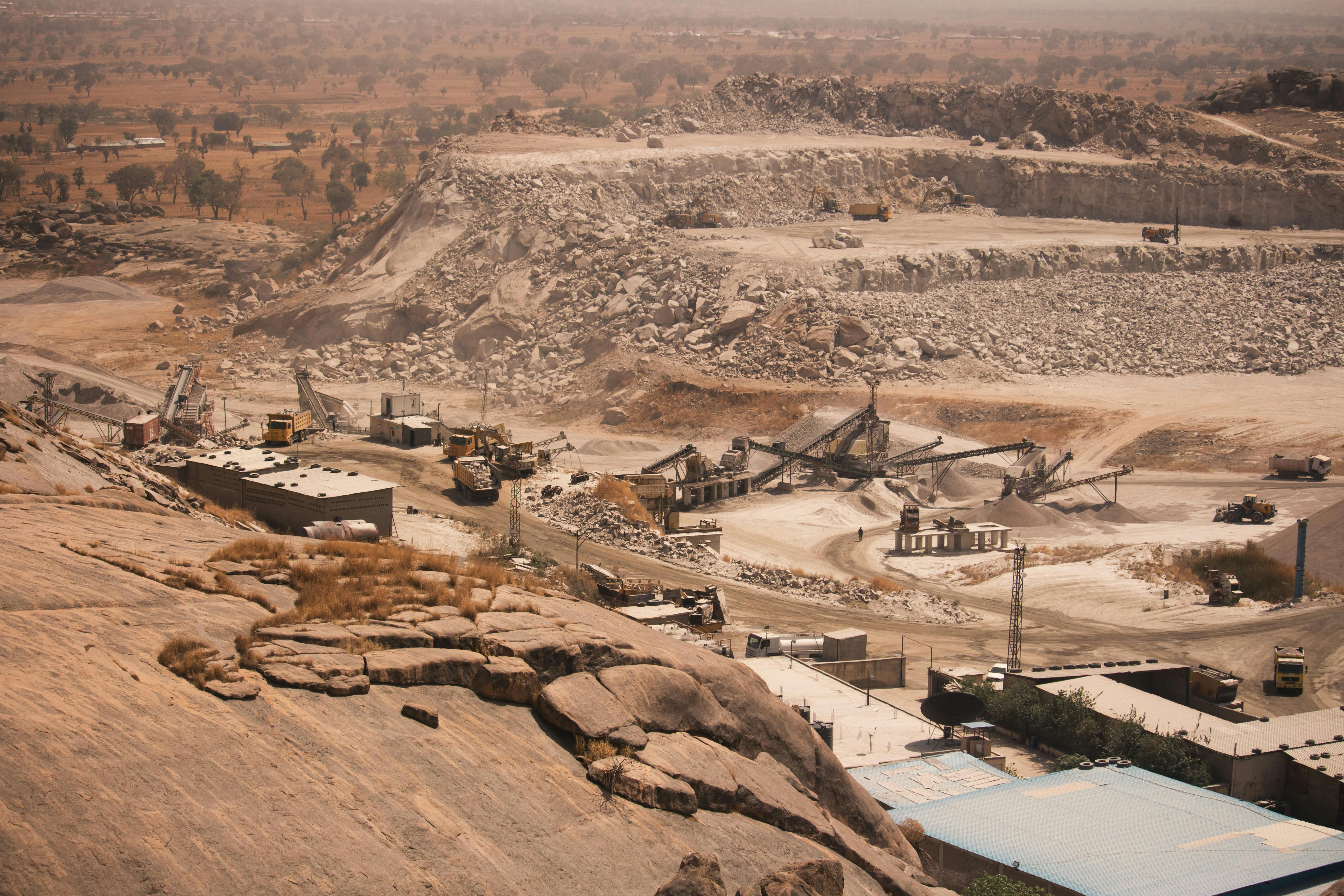 Aerial view of an active stone quarry excavation site with exposed rock faces, extraction equipment, piles of extracted stone material, and a winding river visible in the surrounding landscape under hazy atmospheric conditions.