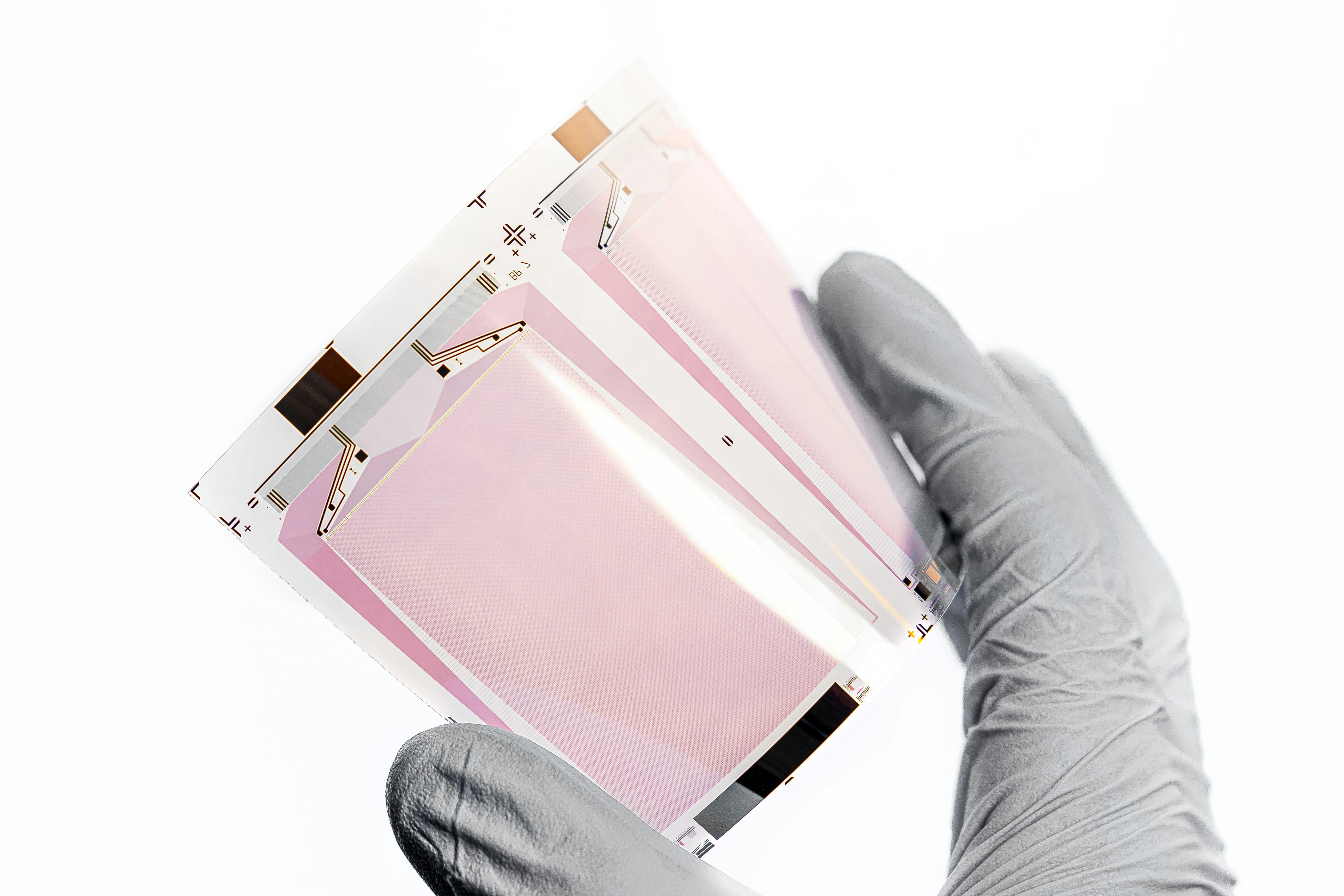 A gloved hand in a laboratory setting holds up a transparent pink battery cell or electrode material sample, displaying its internal layered structure against a white background.