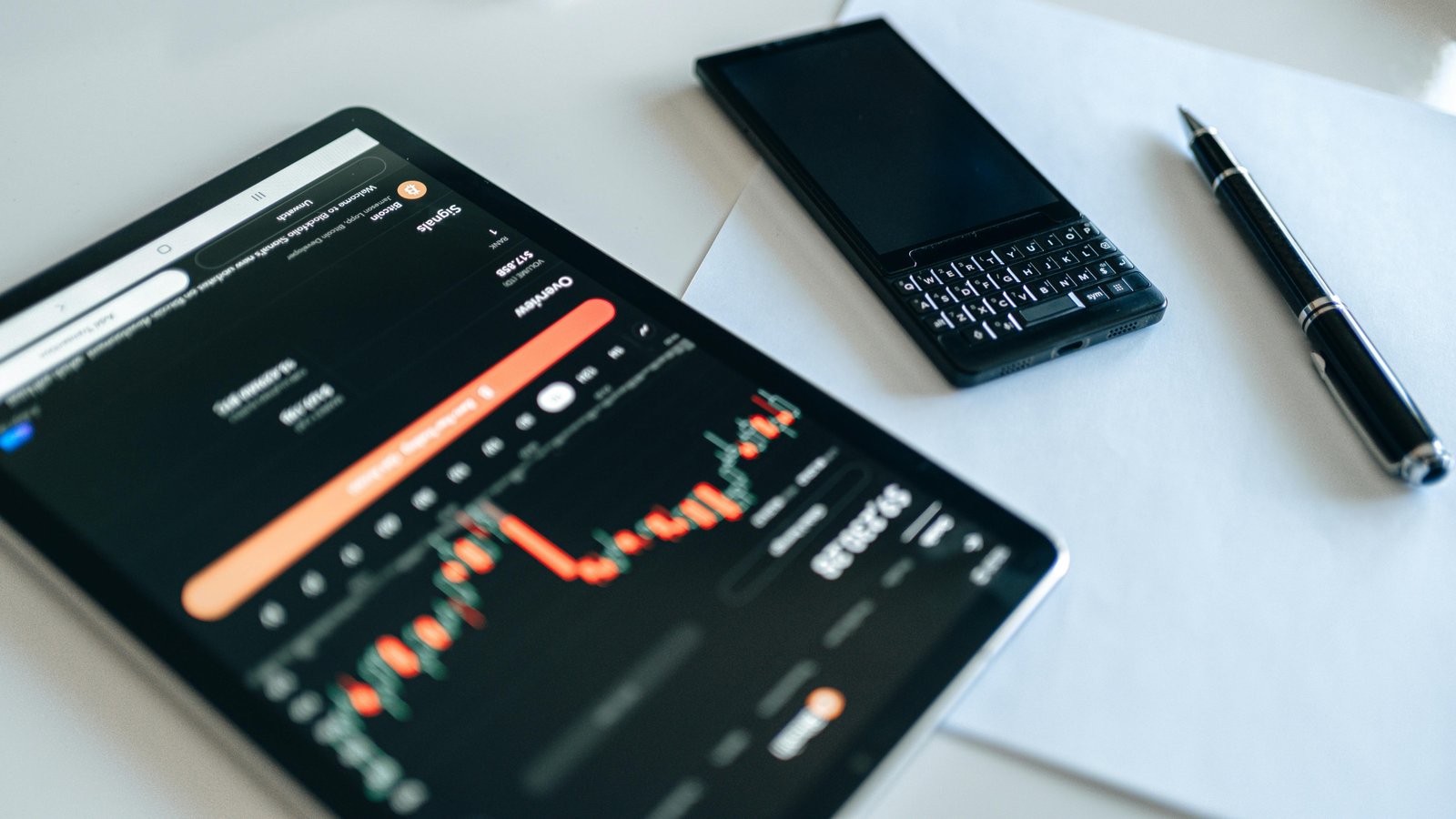 A tablet displaying a dark-themed financial dashboard with orange candlestick charts and data visualization, positioned on a white surface next to a smartphone with a keyboard and a black pen.