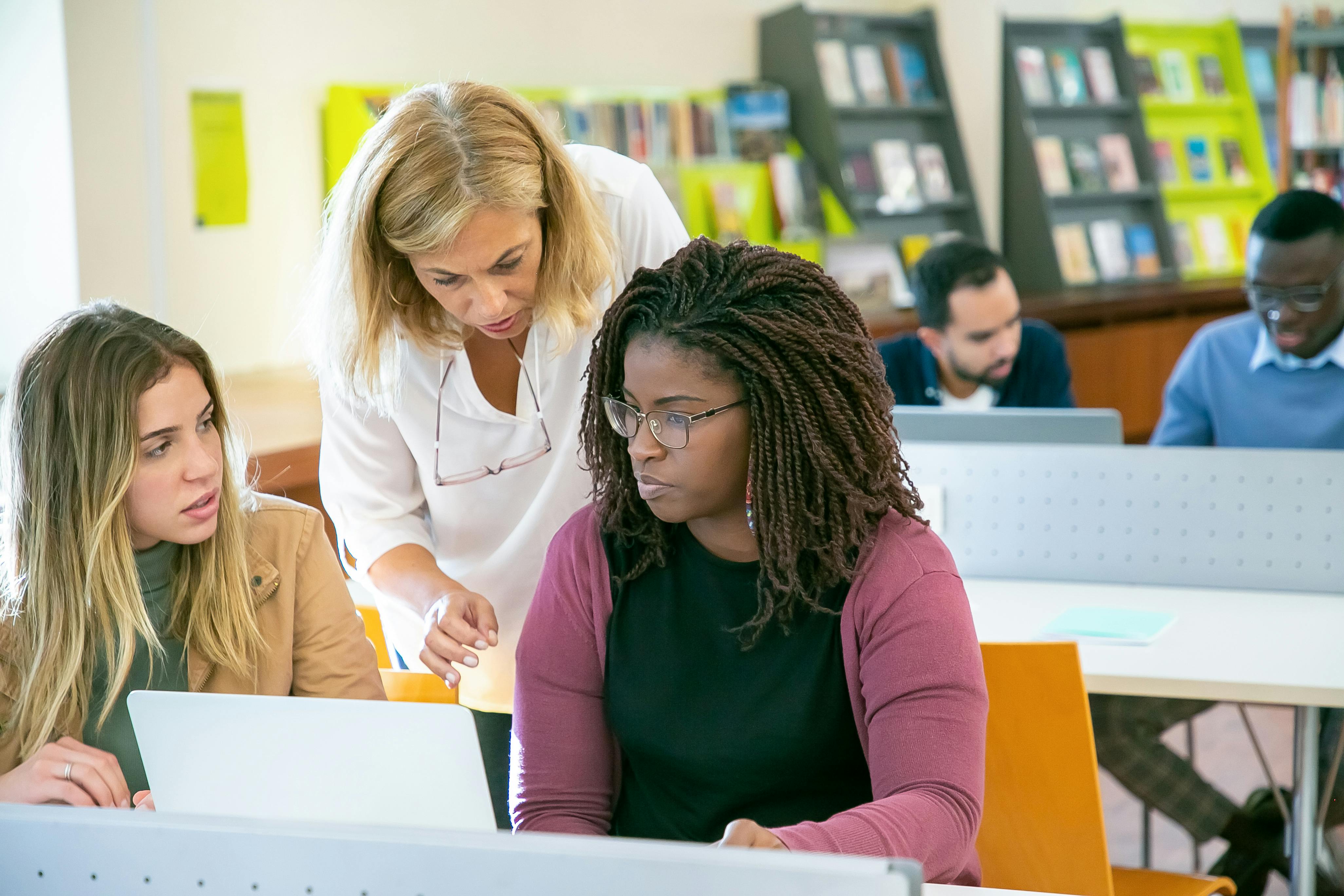 A teacher in a white shirt leans over to assist two female students working at laptops in a bright classroom with colorful shelving and learning materials in the background, while other students work at computers behind them.
