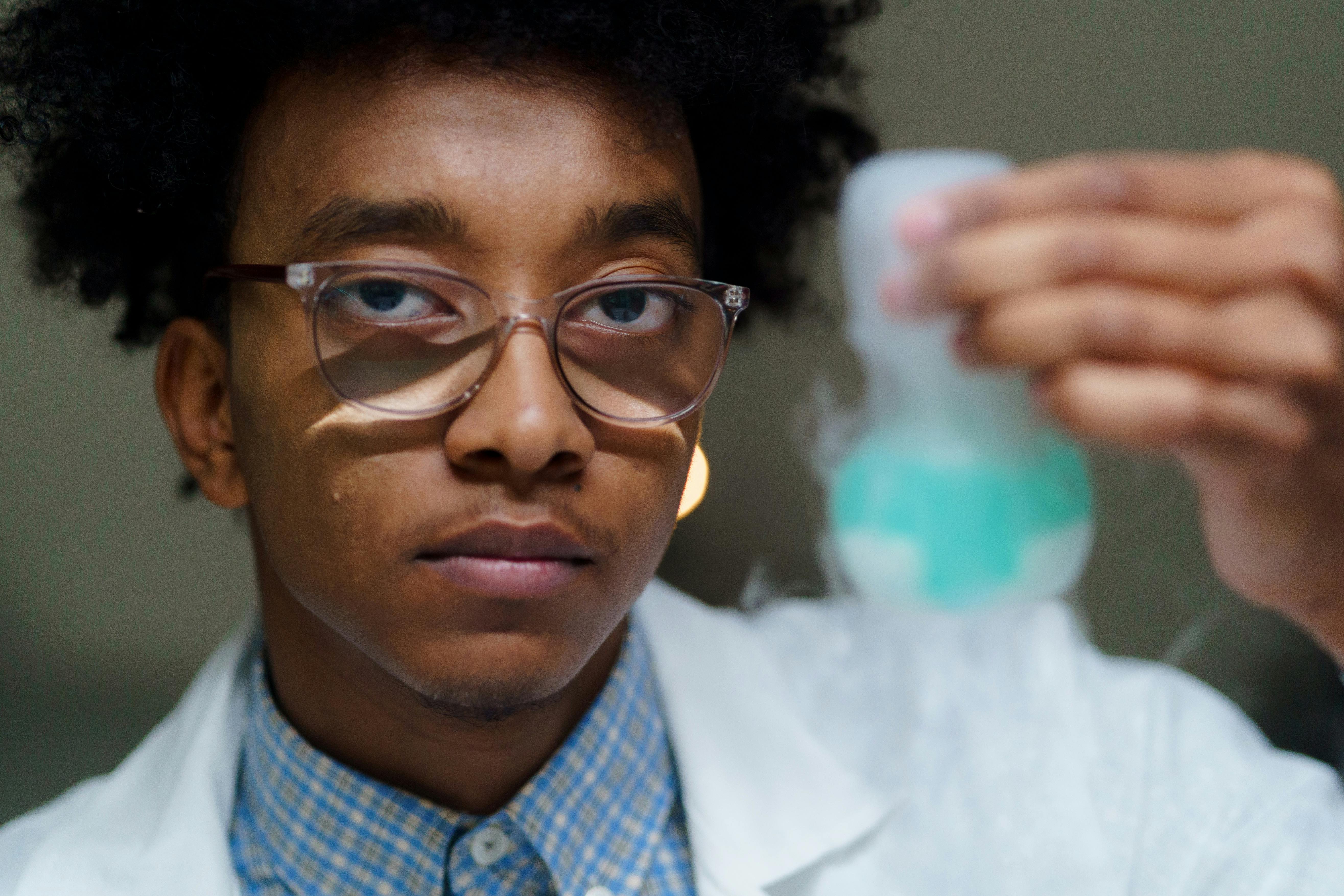 A young person wearing glasses and a white lab coat over a blue checkered shirt examines a turquoise-colored object in their hand, with a blurred hand visible in the background.