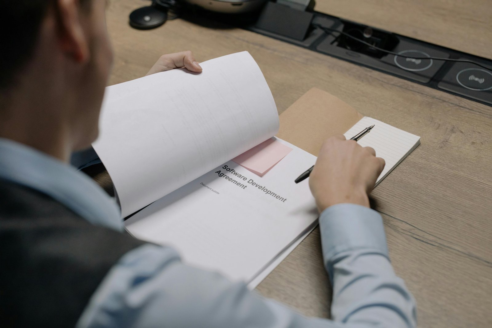 A person in a light blue shirt sits at a wooden desk reviewing printed documents and paperwork with a computer monitor visible in the background.