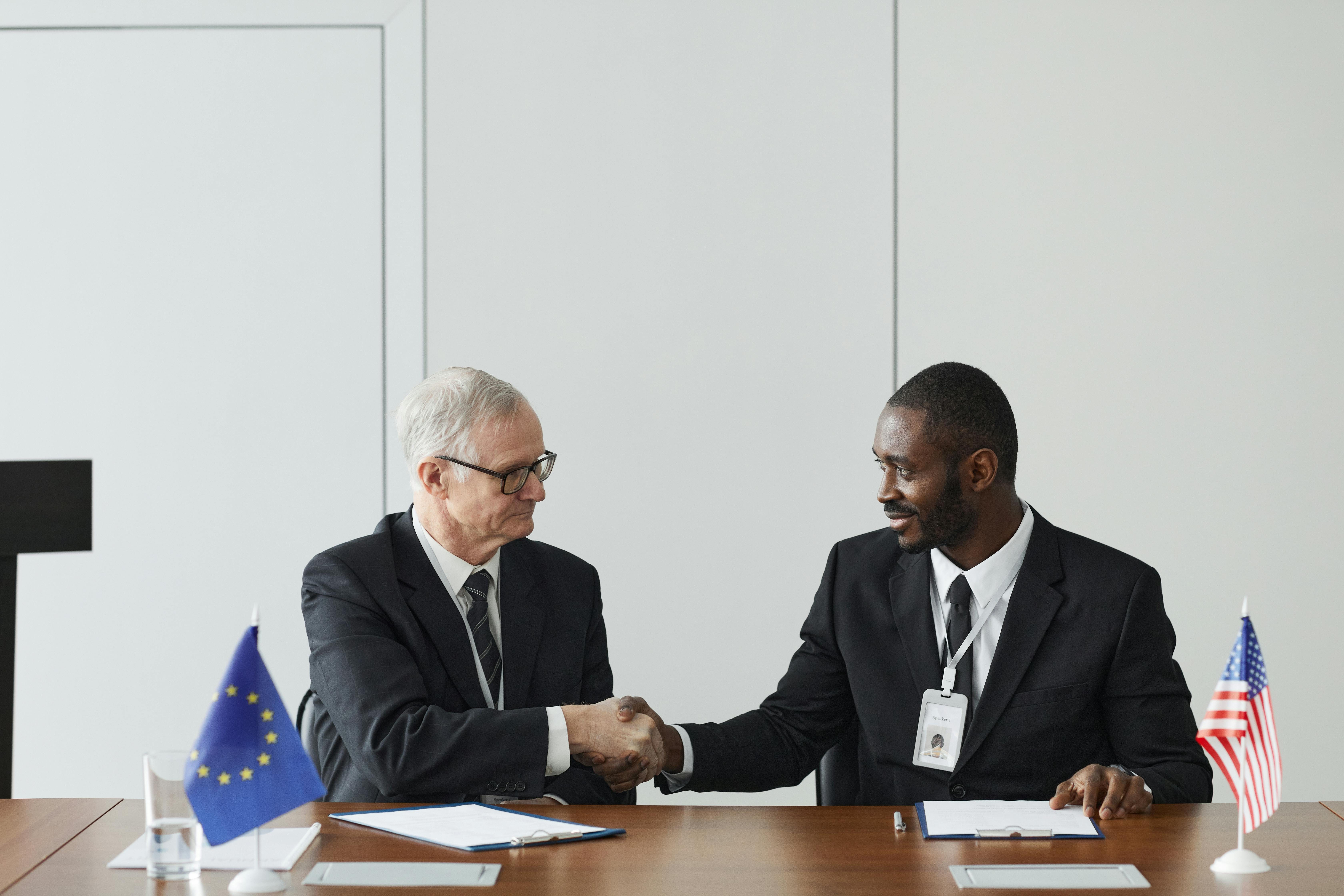 Two professional men in business suits shake hands across a conference table in a formal meeting room, with EU and US flags visible, documents and water glasses on the table, and a neutral white wall backdrop.