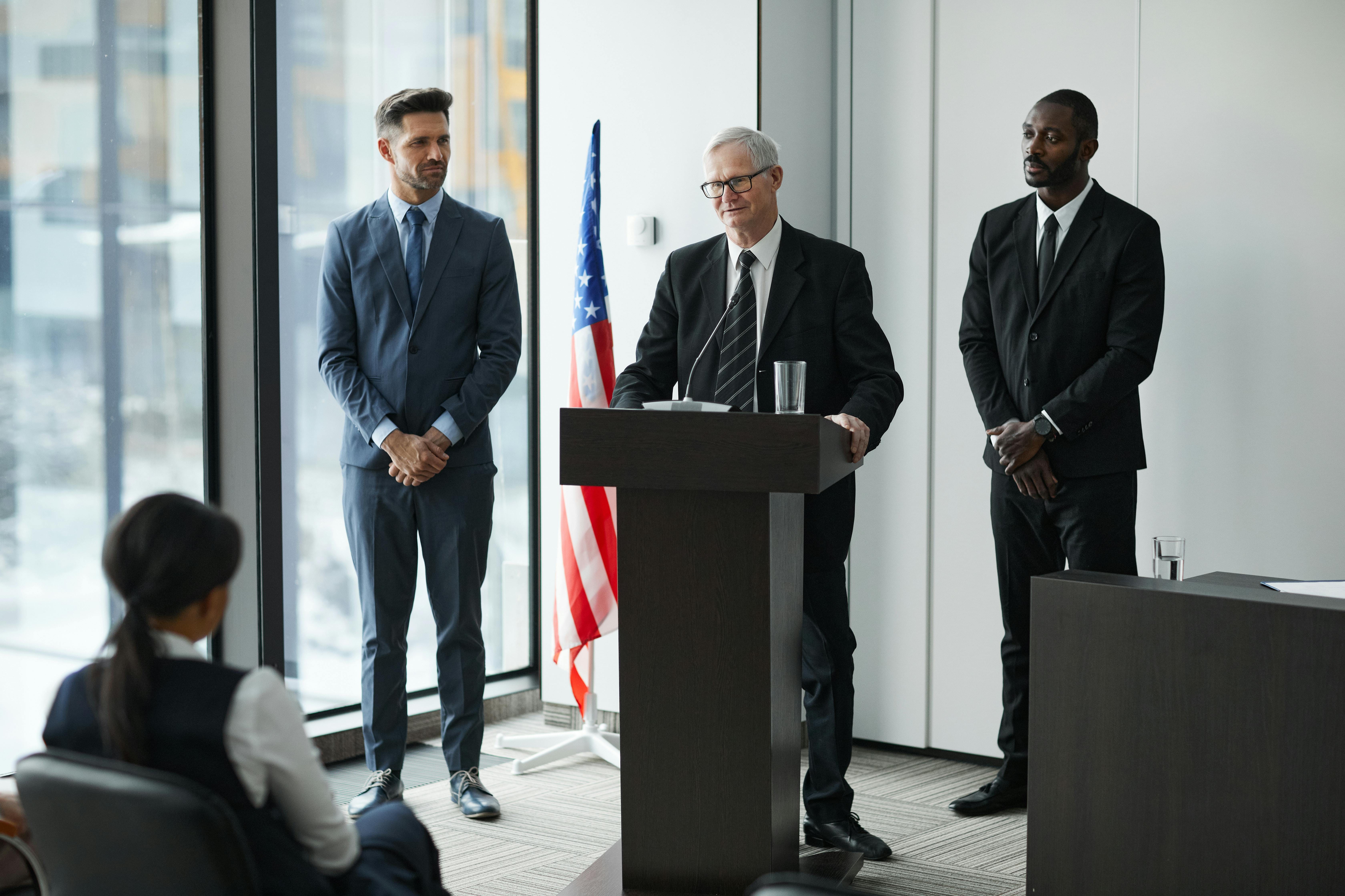 Three men in dark business suits stand in a modern high-rise conference room with floor-to-ceiling windows; a middle-aged man with glasses speaks at a podium bearing an American flag while two other men flank him on either side, with a fourth person visible from behind in the foreground.