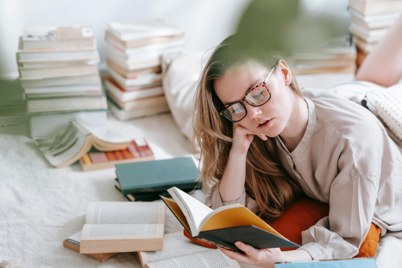 A person wearing glasses lies on a white surface surrounded by stacked books, reading an open book with a contemplative expression while resting their head on their hand.