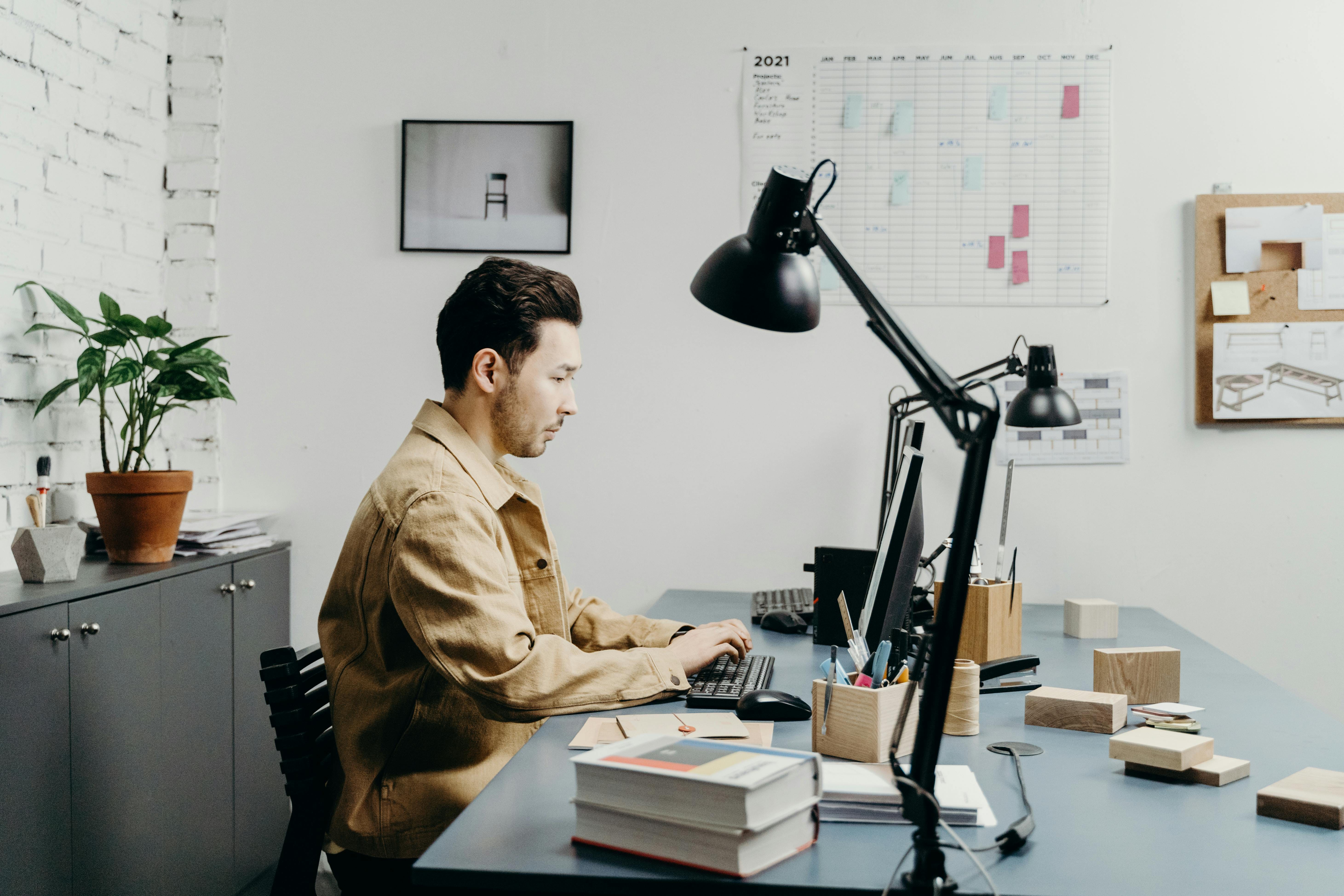 A man in a tan/brown button-up shirt sits at a blue desk in a modern office, typing on a laptop while surrounded by stacked books and papers, with a black adjustable desk lamp, potted plant, wall calendar, and shelving visible in the background.
