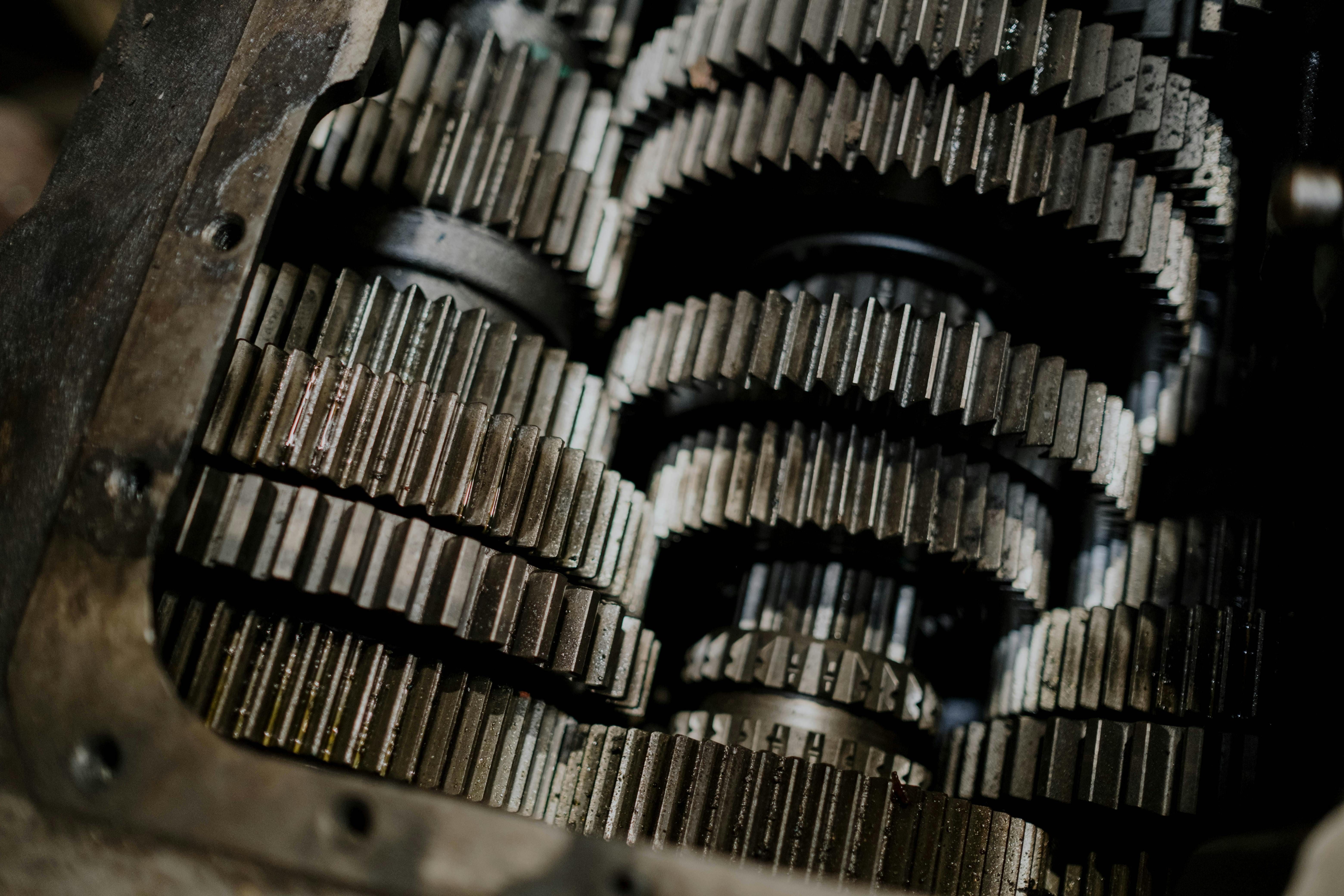 Close-up photograph of multiple large industrial gearbox gears with prominent teeth, showing metal mechanical components in what appears to be a machinery assembly or workshop setting with dramatic lighting.