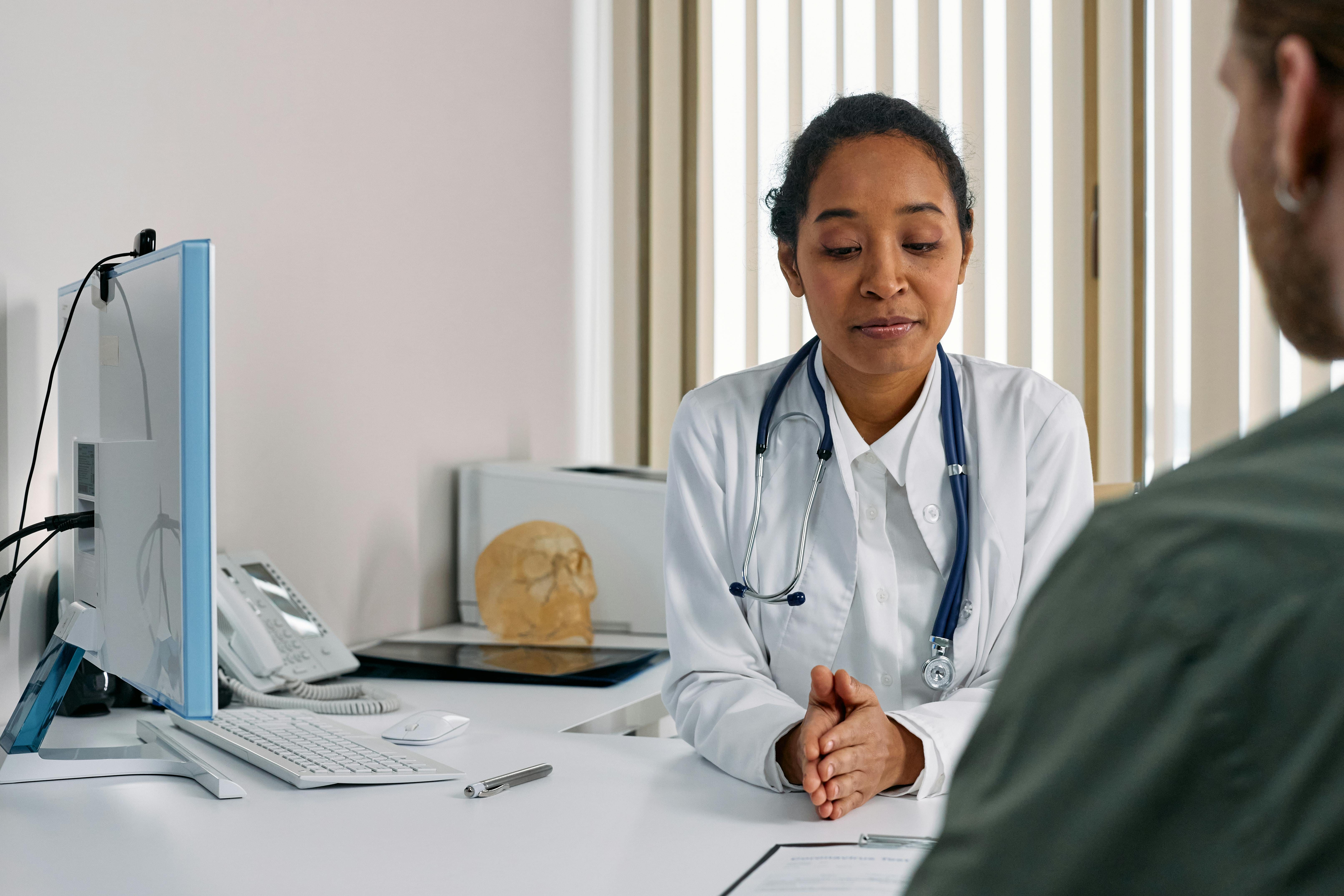 A female healthcare provider in a white coat with a stethoscope sits at a desk across from a male patient in a medical consultation room, with a computer monitor, anatomical model, and office supplies visible.