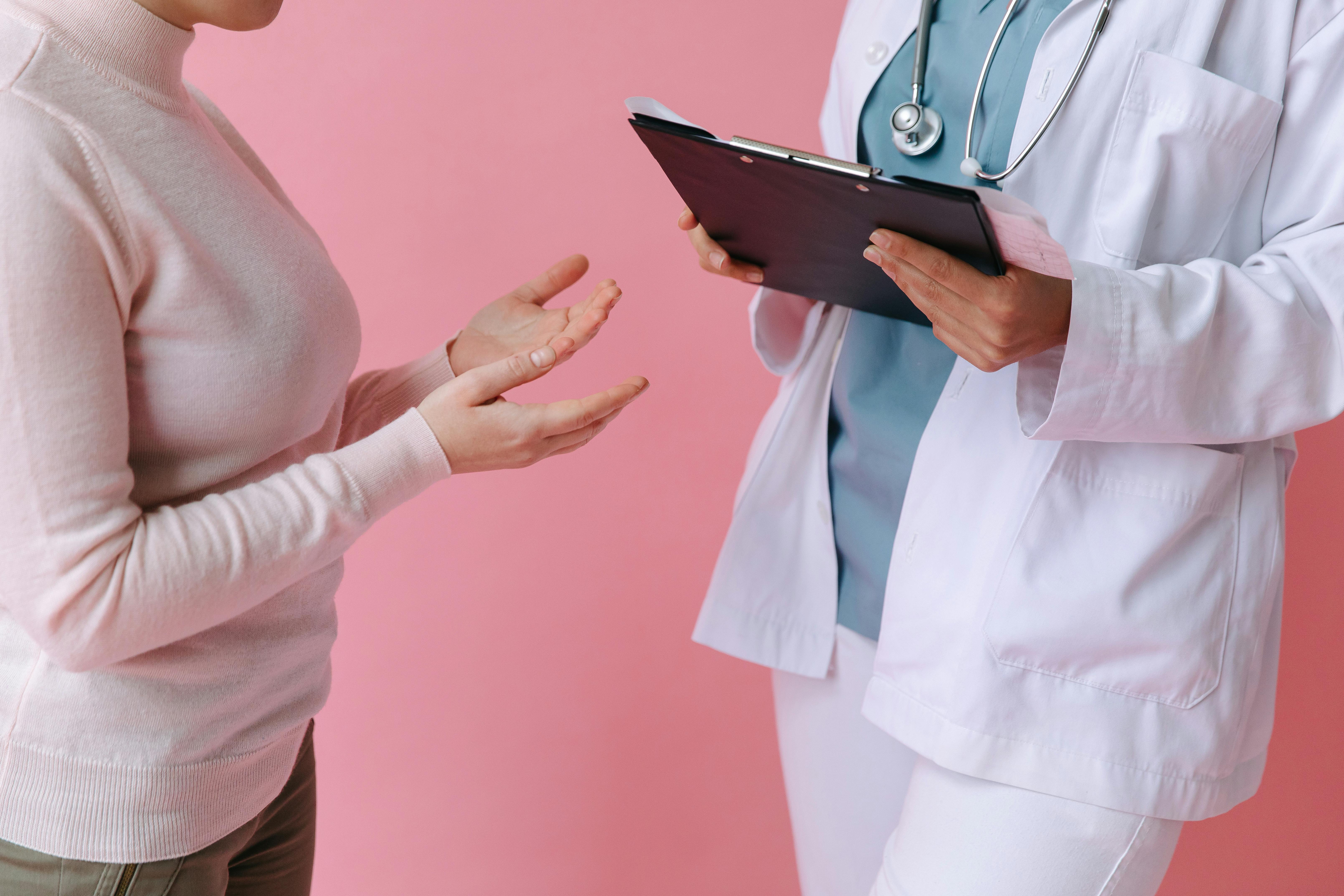 A healthcare provider in a white coat with a stethoscope consults with a pregnant woman in a pink long-sleeved shirt against a pink background, with the provider holding a tablet device.
