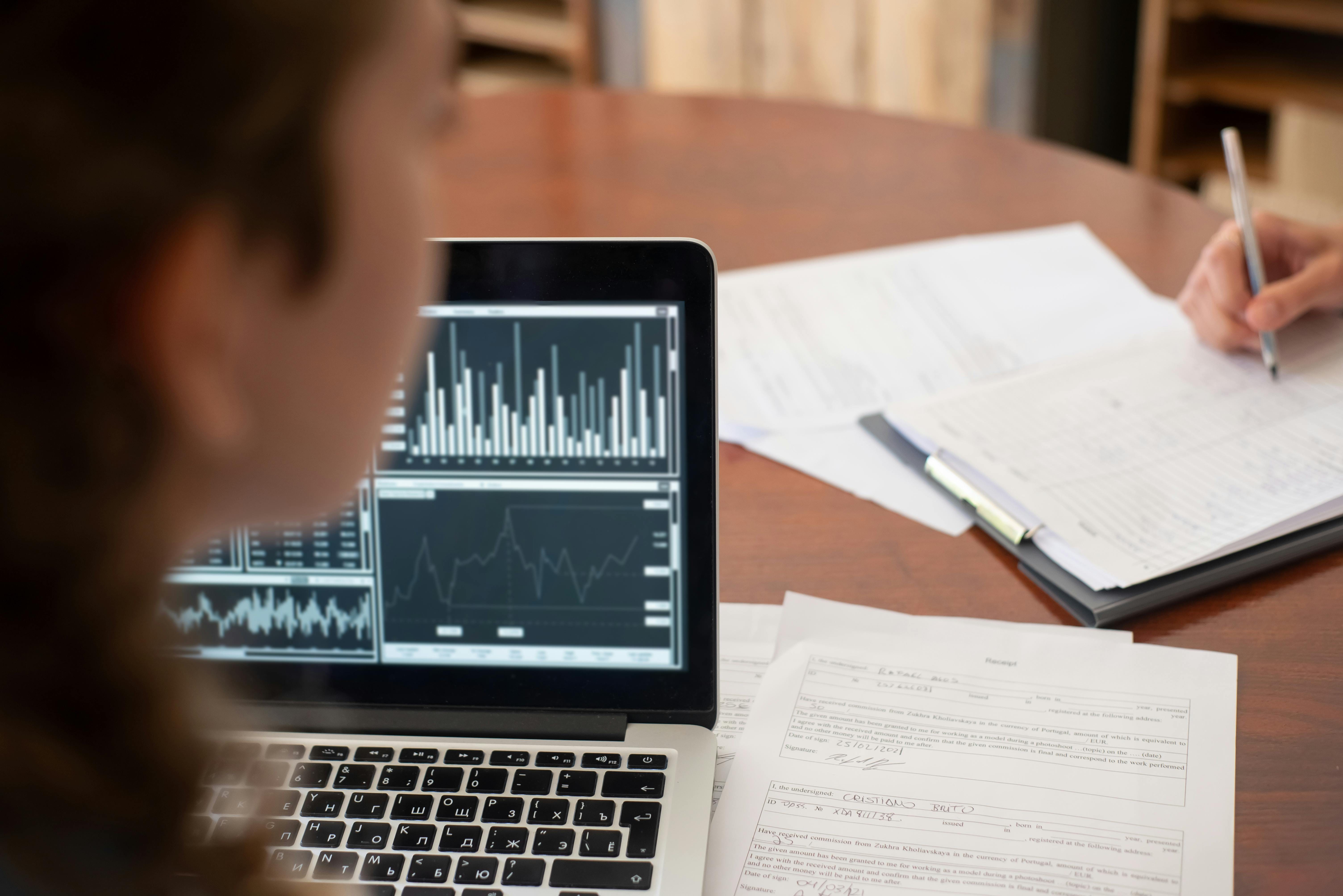 A person seated at a wooden desk works on a laptop displaying data visualization charts and graphs, with printed documents and a notebook visible on the desk surface, suggesting active data analysis work.