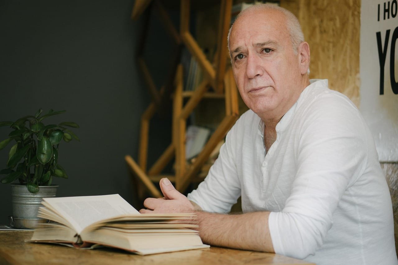 A middle-aged person with blonde hair sits at a wooden desk in a study setting, reading from an open book with a contemplative expression, with additional books visible on the desk and warm natural lighting coming from the left.