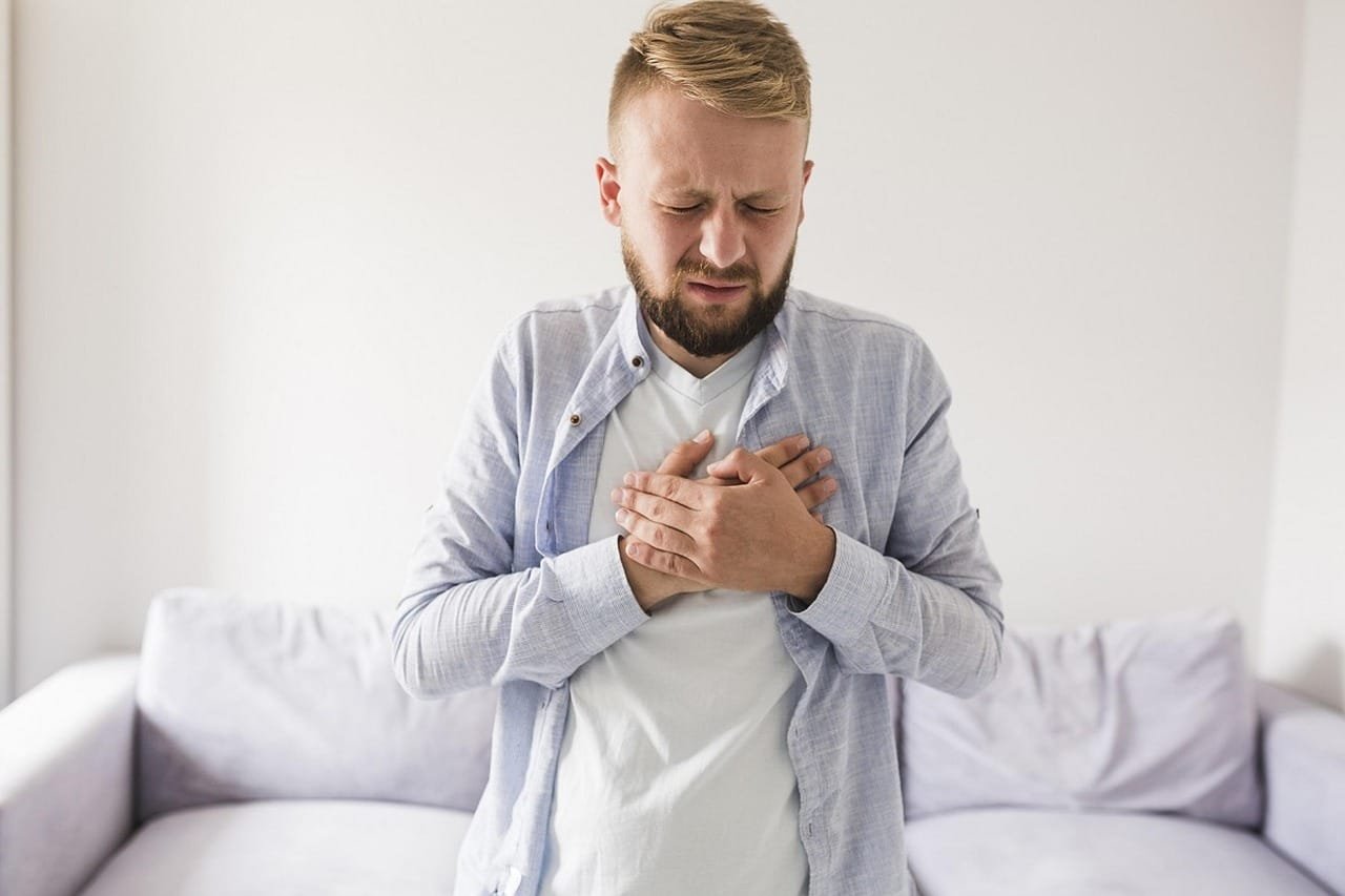 A young man in a light blue shirt sits on a bed holding his chest with both hands, appearing to experience chest discomfort, while a healthcare professional in a white coat stands beside him in a clinical setting.