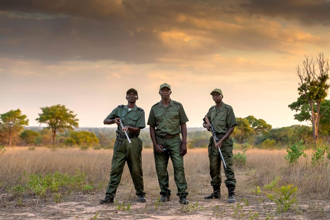 Four uniformed officers in tactical gear stand in a line across a dusty landscape at sunset or sunrise, suggesting a border or patrol checkpoint area.