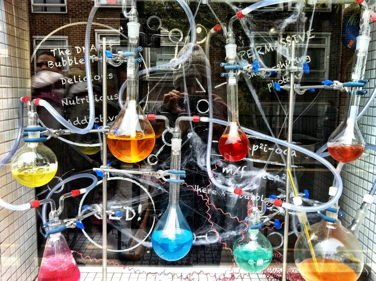A close-up overhead view of a chemistry laboratory setup featuring multiple glass flasks, beakers, and round-bottom flasks containing colorful liquids in shades of yellow, orange, pink, blue, and purple, arranged on a dark surface with laboratory equipment and molecular models visible in the background.