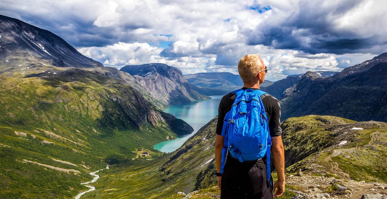 A person wearing a bright blue jacket stands on a mountain ridge overlooking a vast alpine valley with green meadows, scattered farmland patches, and distant peaks under a partly cloudy sky.
