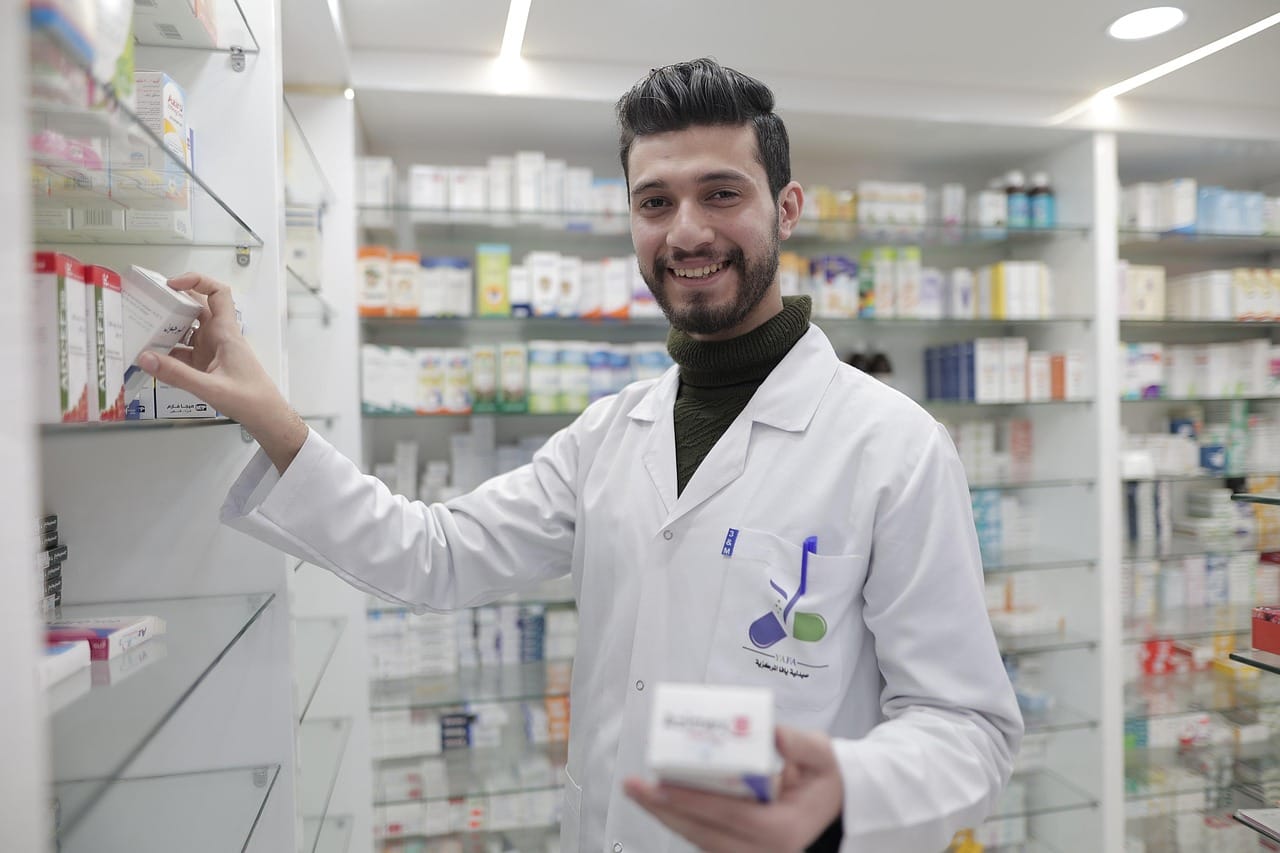 A male pharmacist in a white coat stands behind a pharmacy counter holding a medicine bottle, with shelves of pharmaceutical products visible in the background.