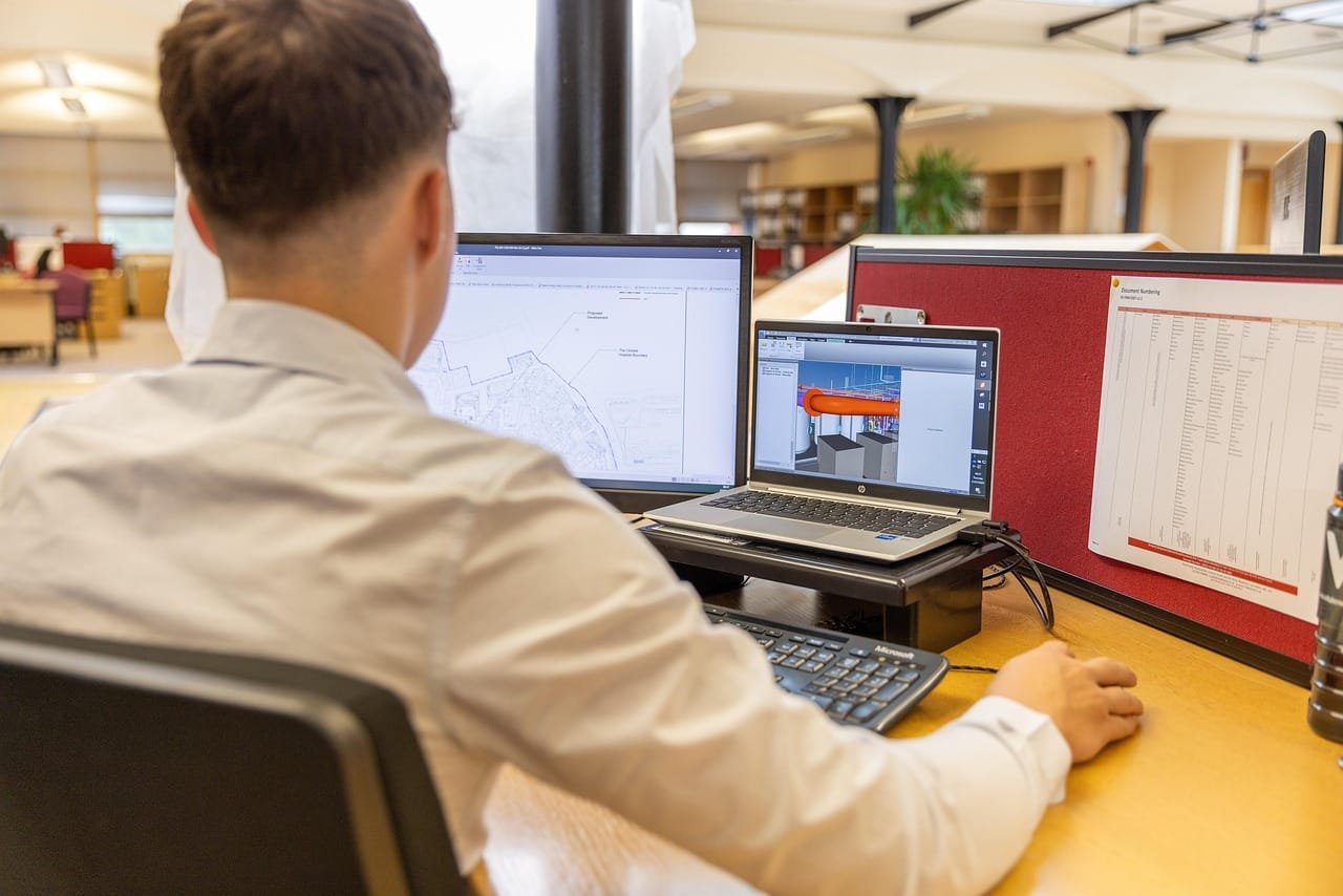 A person seated at a desk working at a computer with a monitor displaying code or development software, in a casual indoor workspace with wooden flooring and furniture visible in the background.