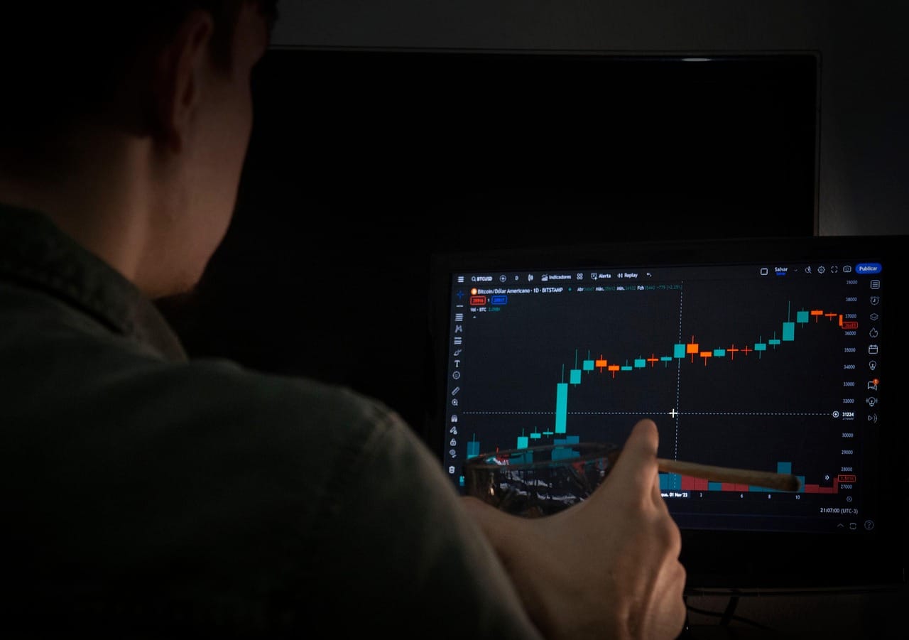 A person viewed from behind sits at a desk in dim lighting, looking at a digital display screen showing blue financial charts and data visualizations on what appears to be a computer monitor.