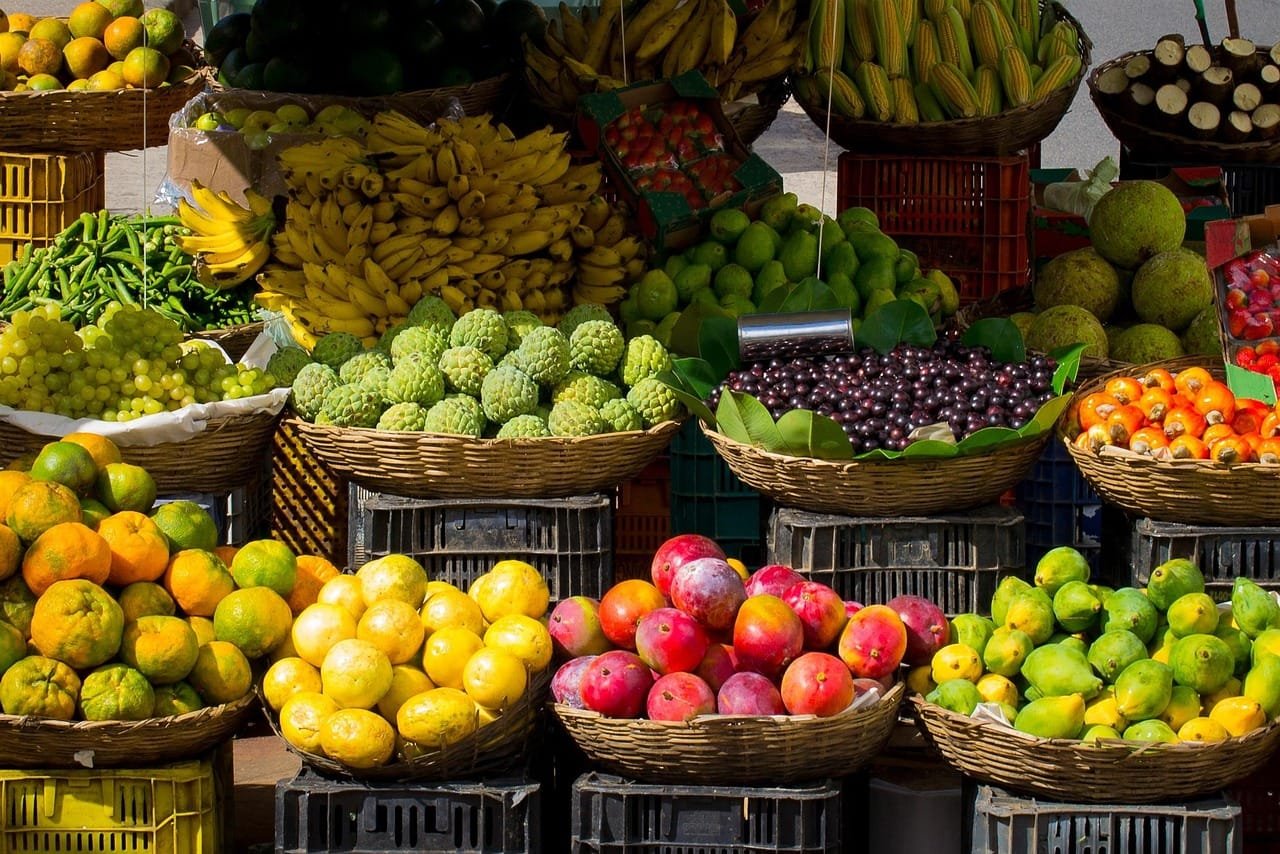 A vibrant outdoor farmers market display featuring colorful fresh produce including yellow lemons, oranges, red apples, limes, and various other fresh vegetables and fruits arranged in wooden crates and containers.