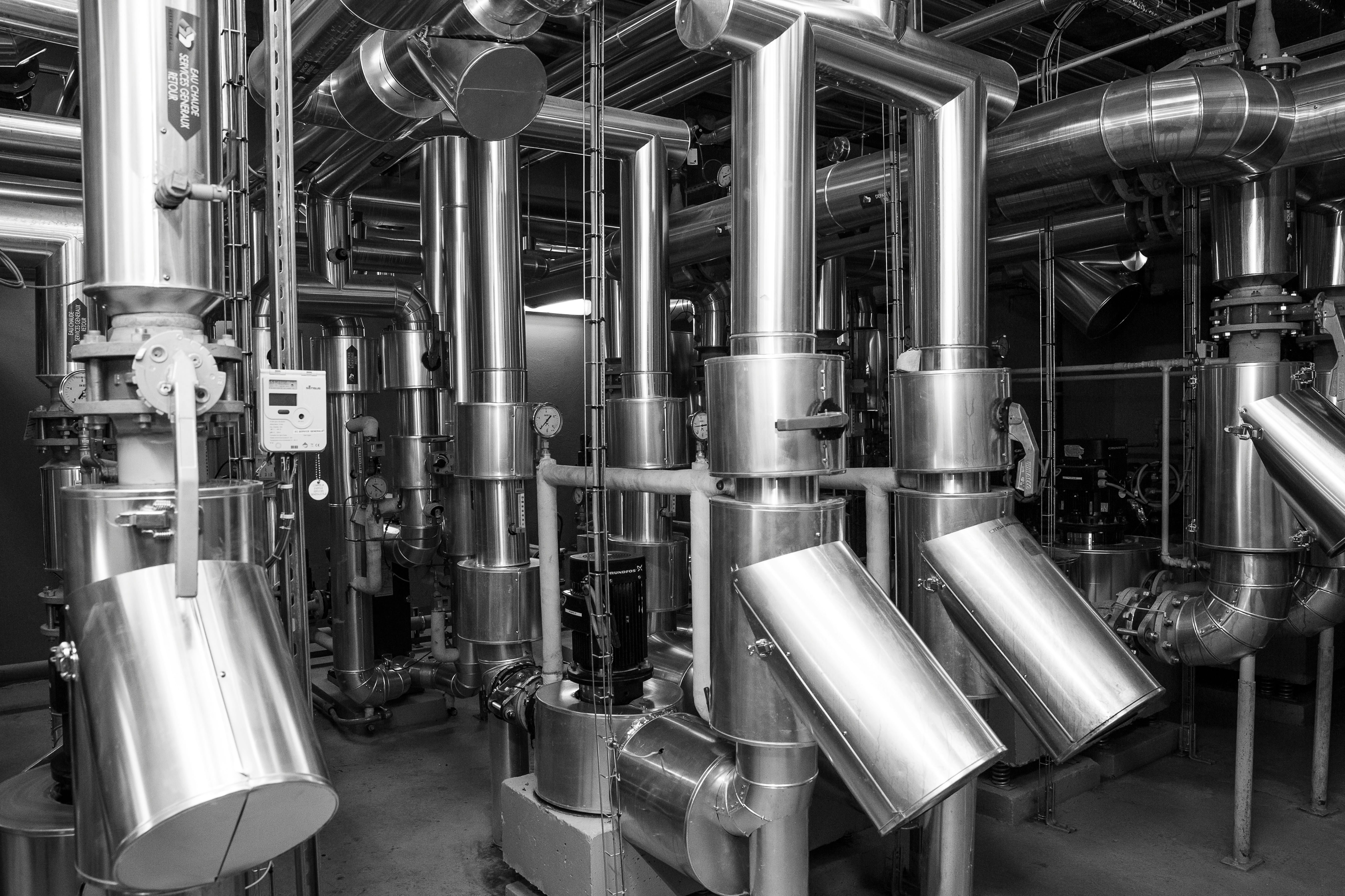 A black and white photograph of an industrial facility interior showing an extensive network of polished metal pipes, cylindrical vessels, and ductwork arranged vertically and horizontally, typical of a research or nuclear facility's thermal management system.