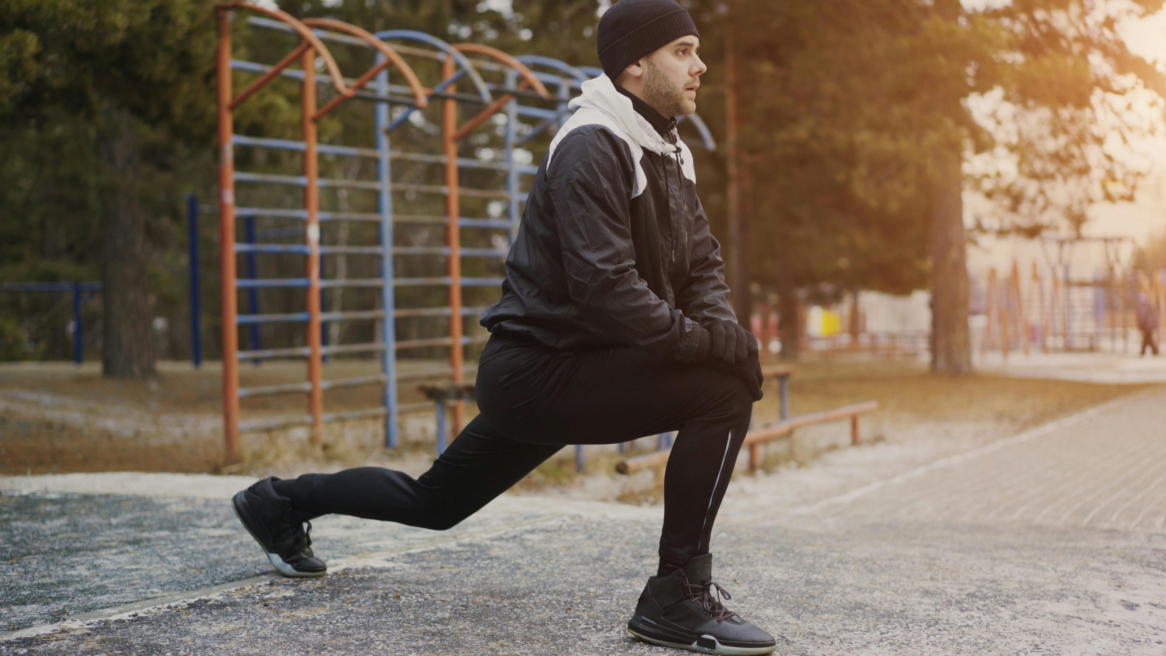A person in dark athletic clothing performs a lunge stretch outdoors on a paved path during golden hour, with metal exercise bars and trees visible in the background.