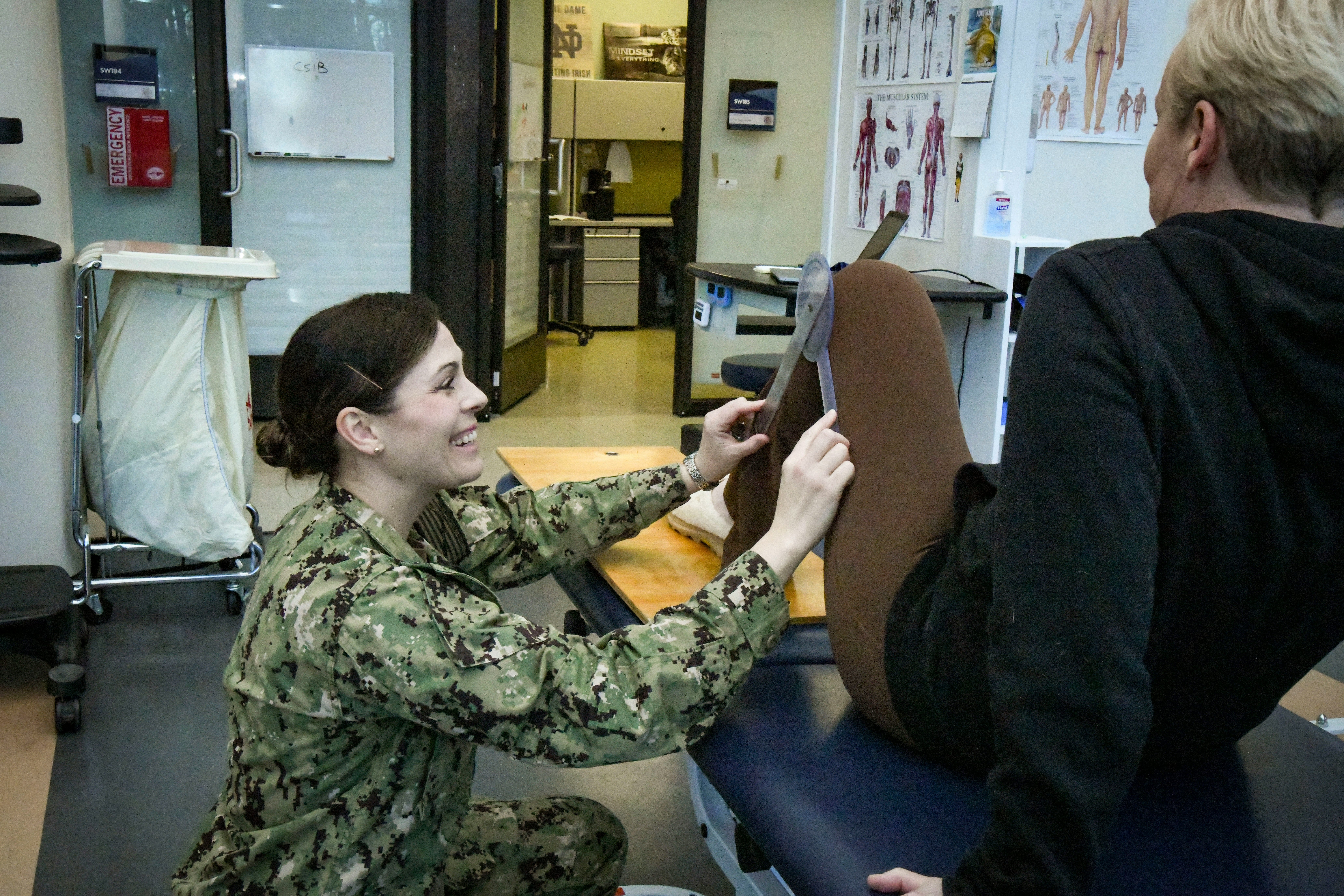 A person in a naval uniform sits at a desk facing another person, reviewing documents together in an indoor office setting with papers and notices visible on the walls and shelves in the background.