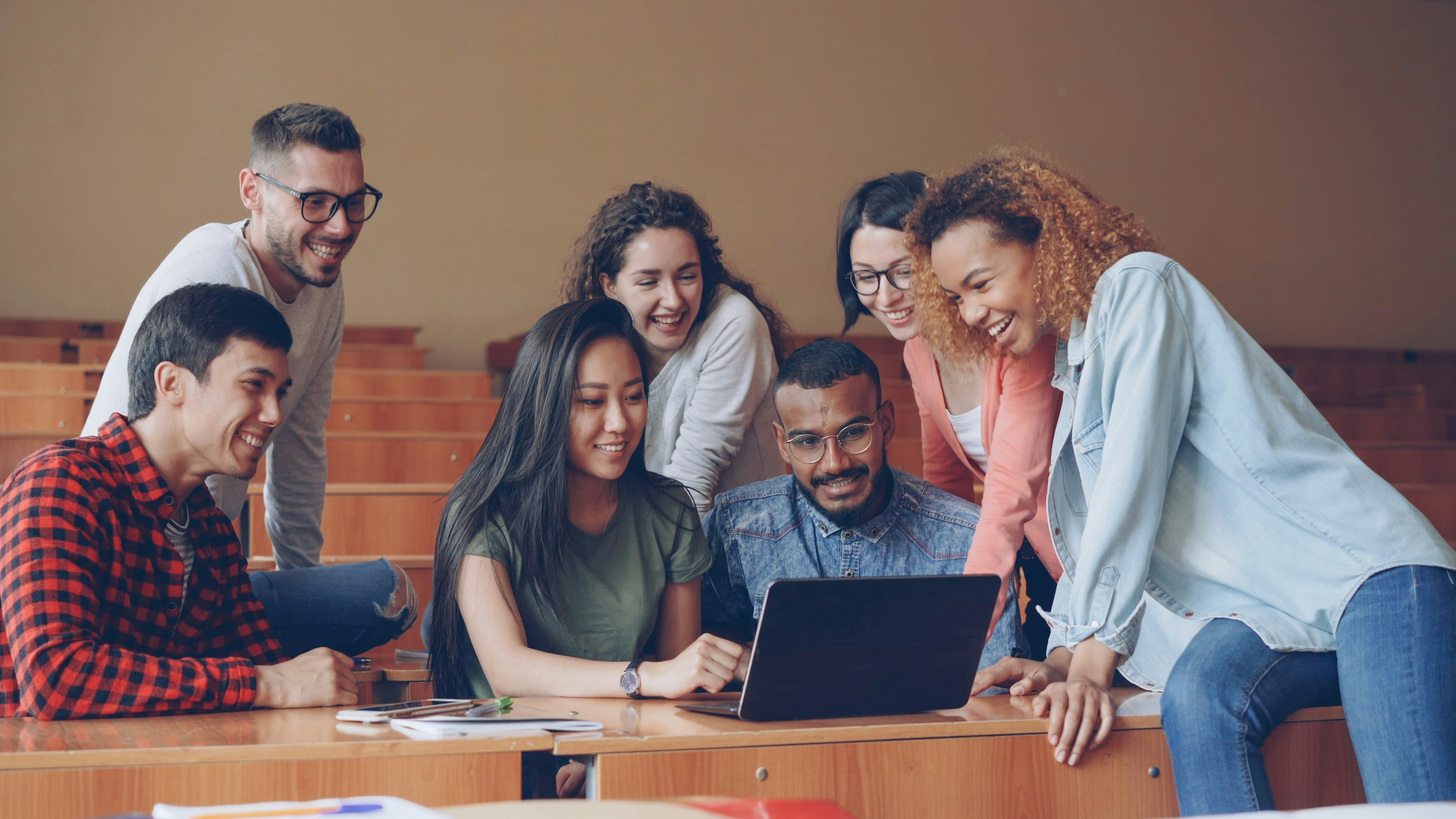 A diverse group of seven people of various ages and ethnicities gathered around a wooden table, smiling and looking at a laptop together in what appears to be a community or collaborative meeting space with warm lighting.