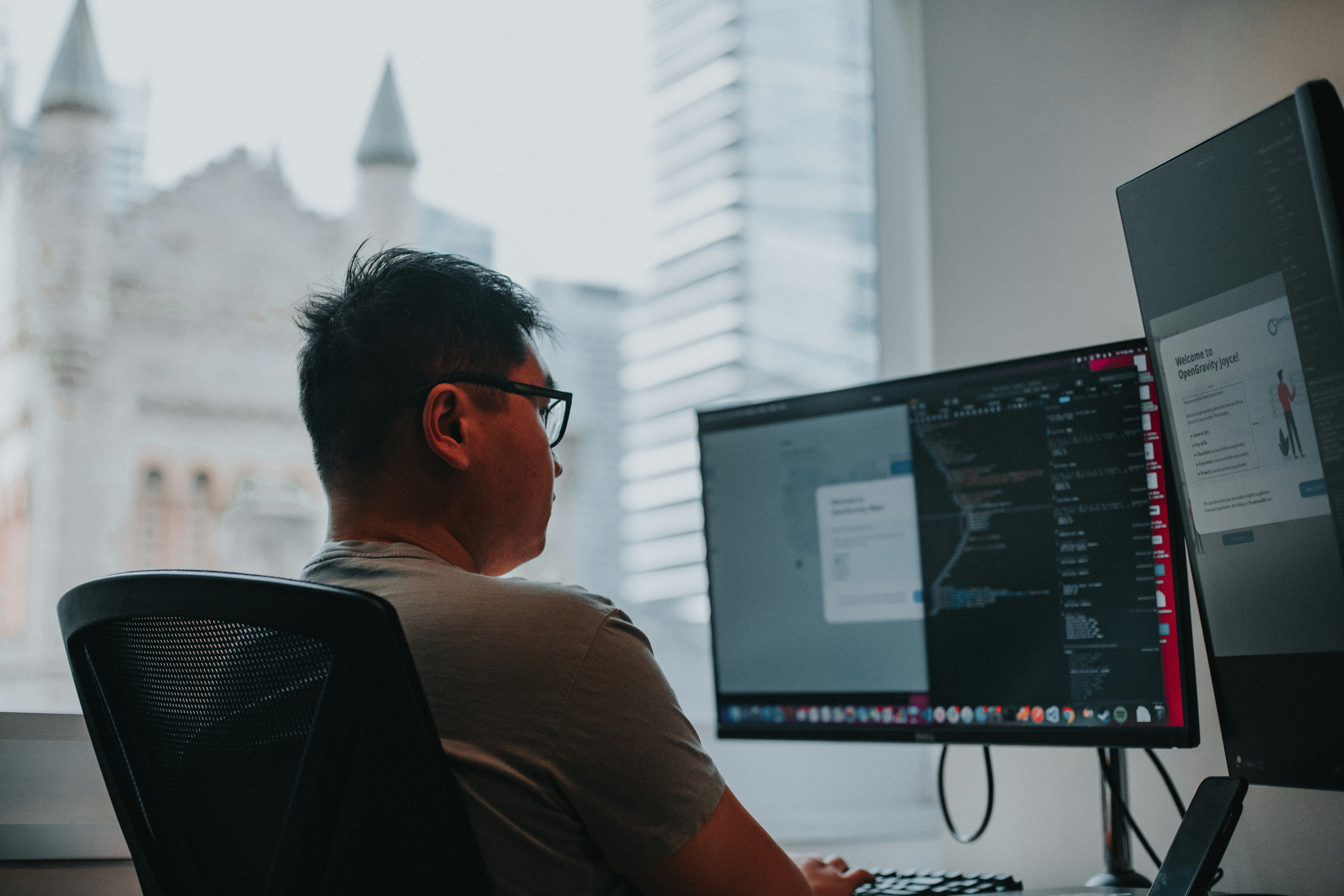 A person wearing glasses sits at a desk in a modern office with city skyline visible through windows, viewing multiple computer monitors displaying code and digital interfaces.