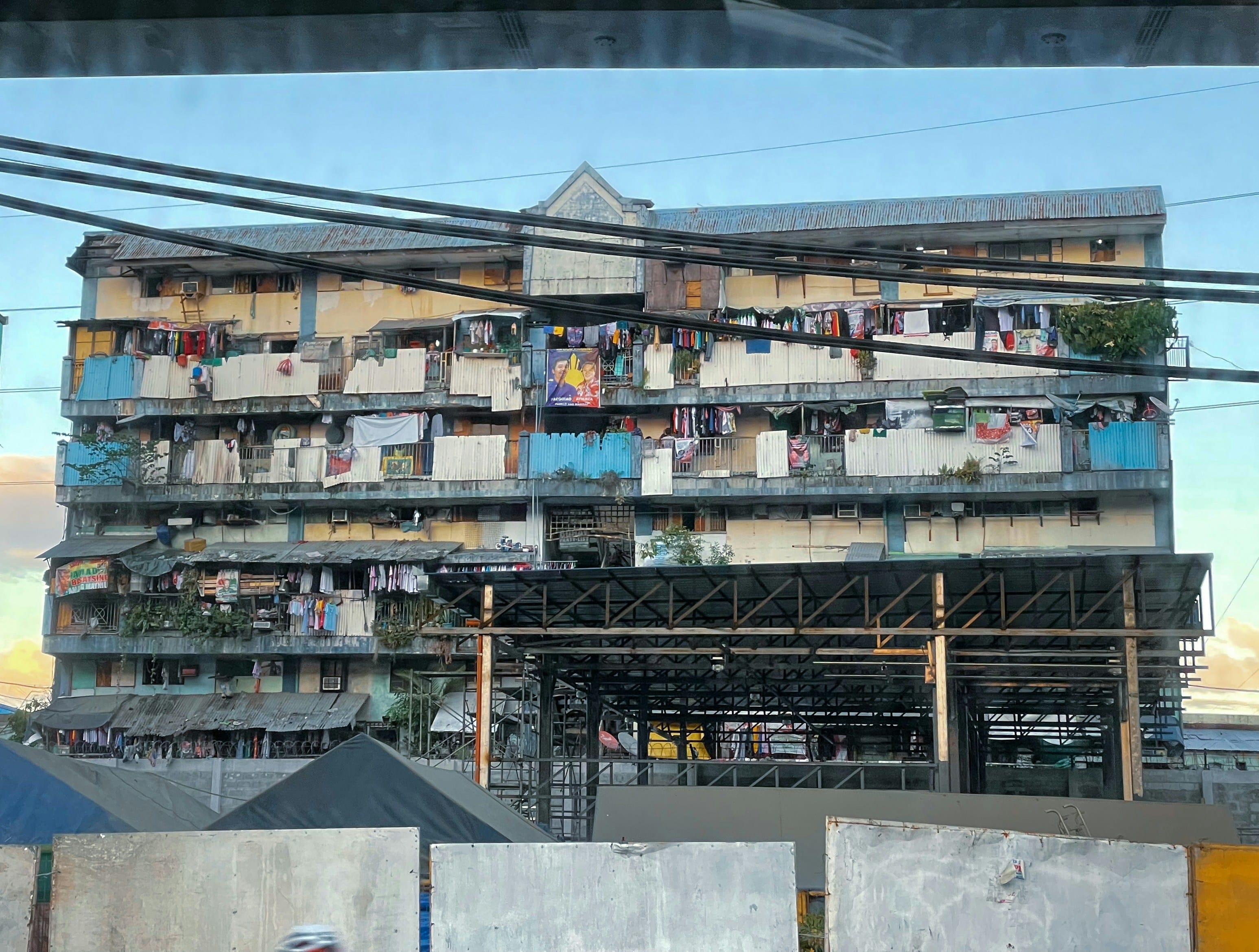 A densely packed multi-story informal residential building in an urban Asian setting, with makeshift structures, blue tarps, laundry lines, and weathered facades showing crowded urban living conditions.