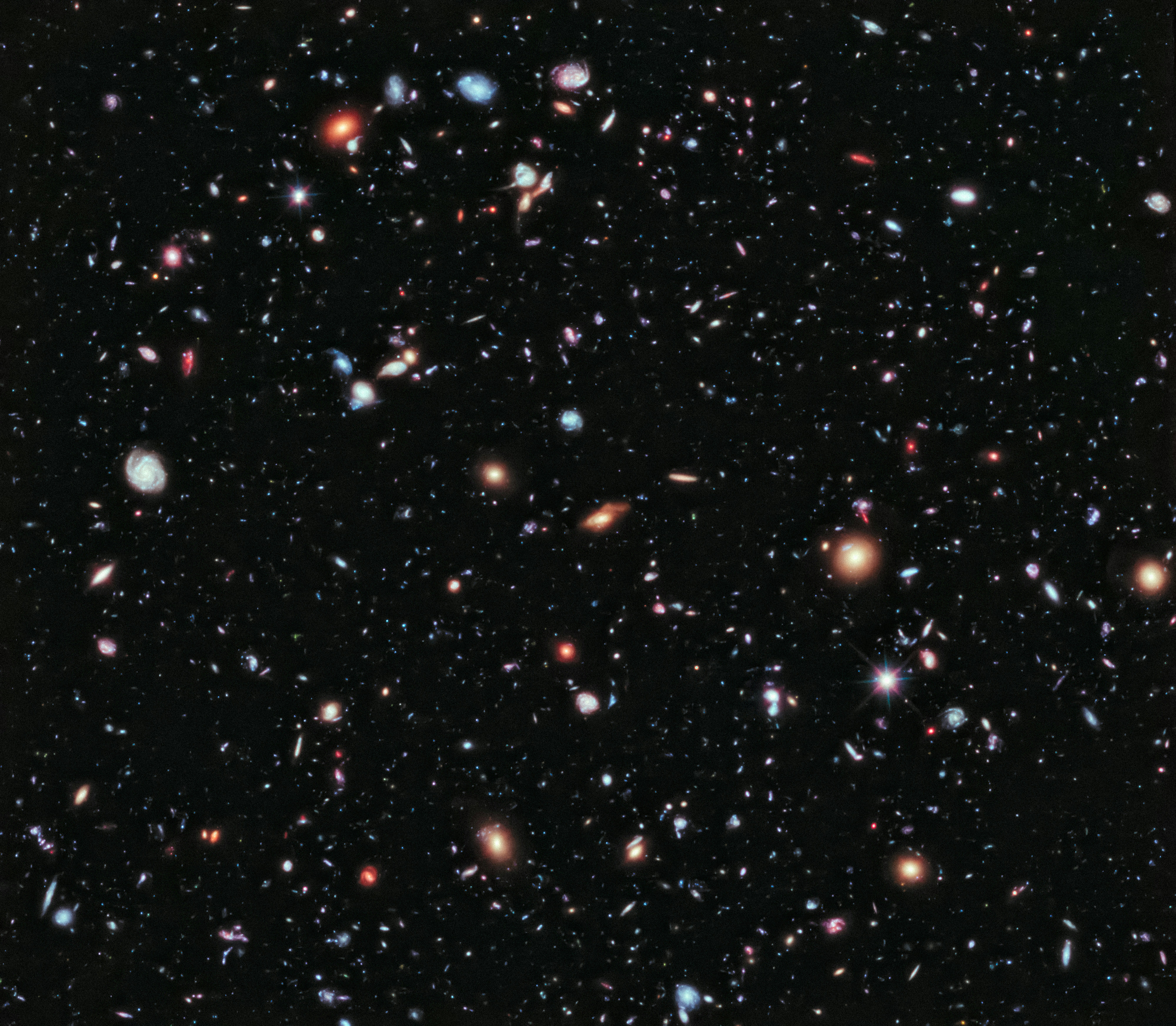 A deep space astronomical field showing hundreds of distant galaxies and stars of varying colors and brightness scattered across a black cosmos, captured as a wide-angle view of the distant universe.