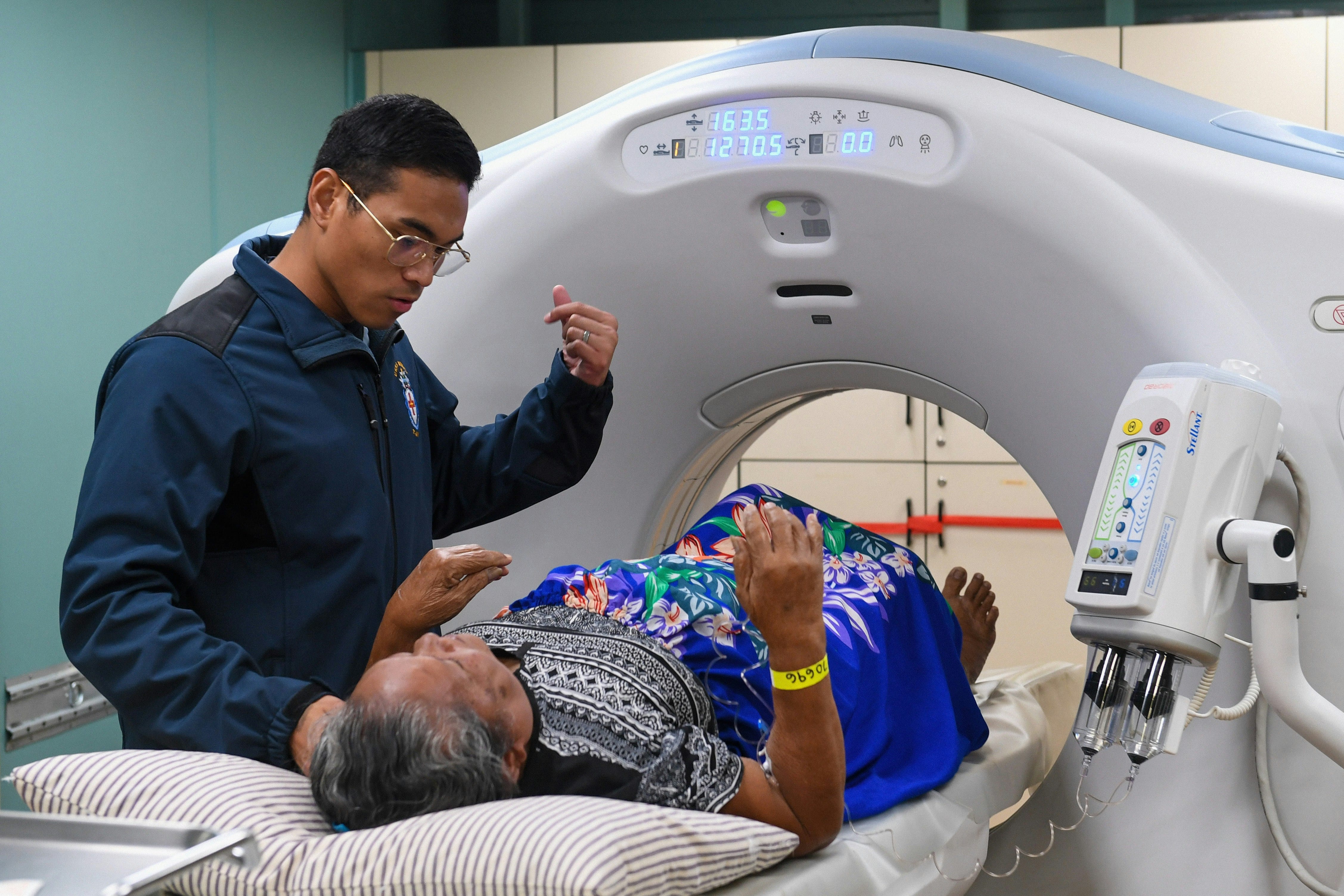 A healthcare professional in a dark blue jacket operates or monitors a modern MRI scanner while a patient lies inside the machine in a medical imaging facility.