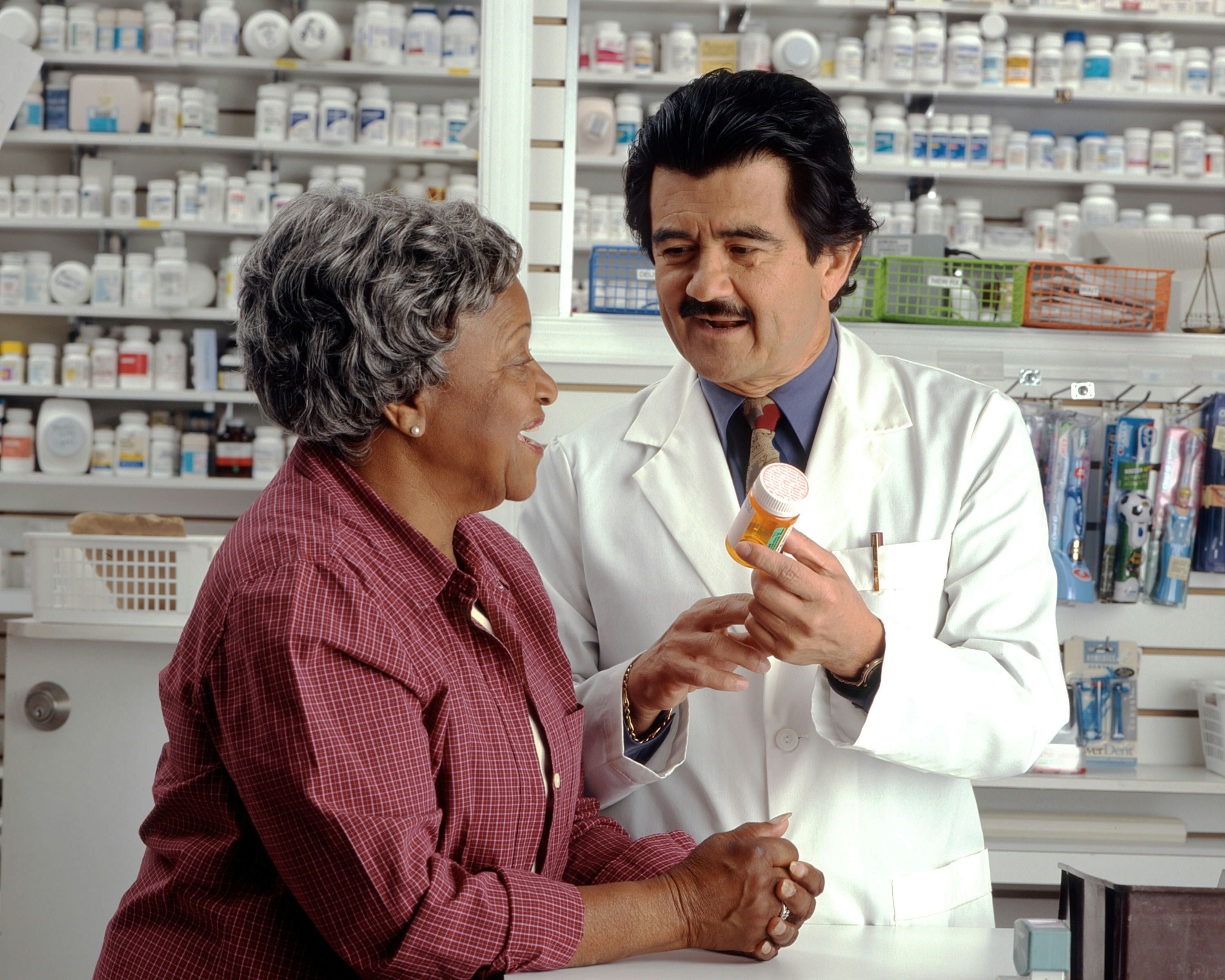 A pharmacist in a white coat speaks with an elderly woman in a burgundy shirt at a community pharmacy counter, with medication shelves visible in the background.