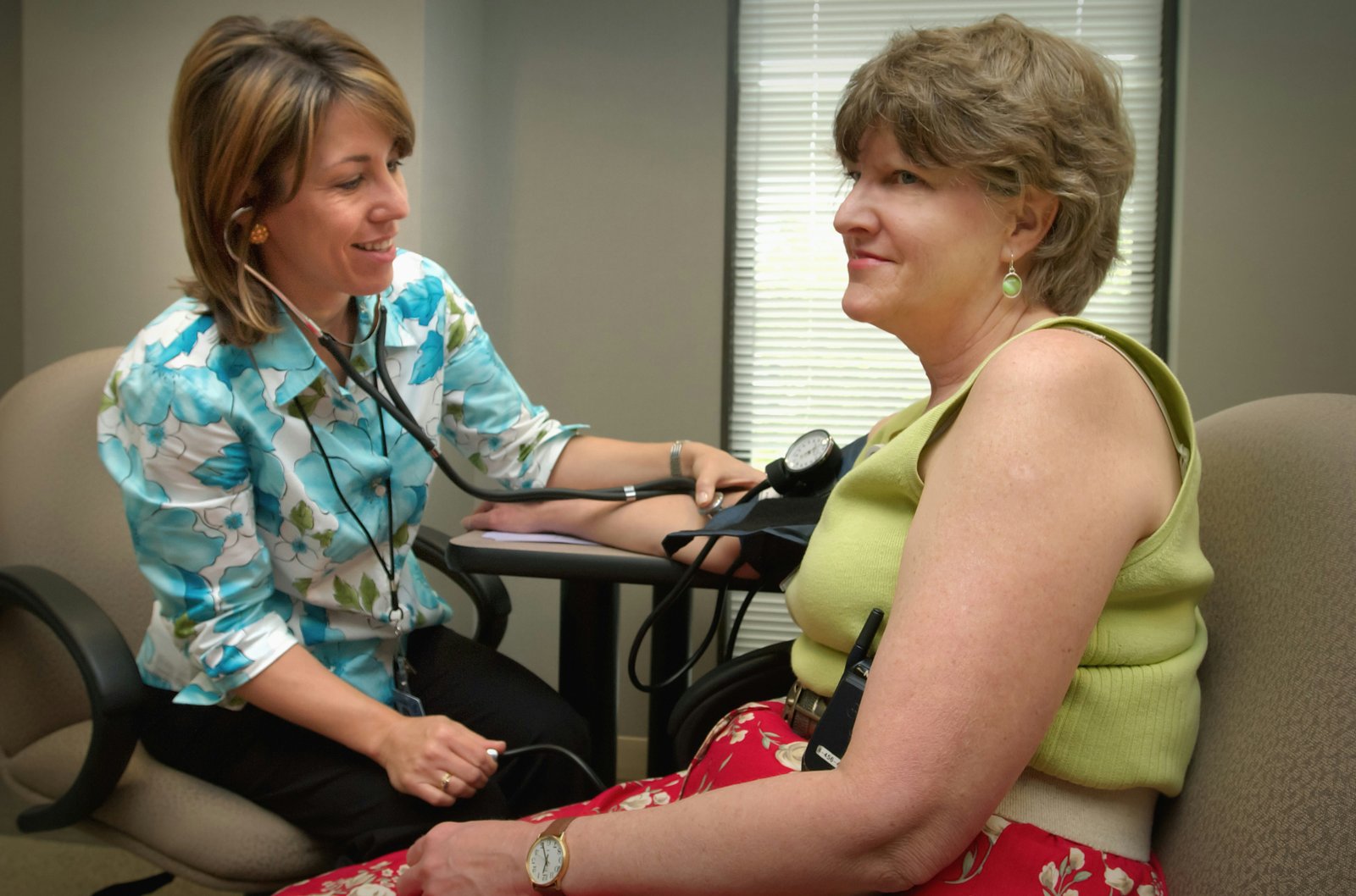 A healthcare provider wearing a blue and teal patterned shirt takes a blood pressure reading from a middle-aged woman wearing a lime green sleeveless top in a clinical office setting with blinds visible in the background.