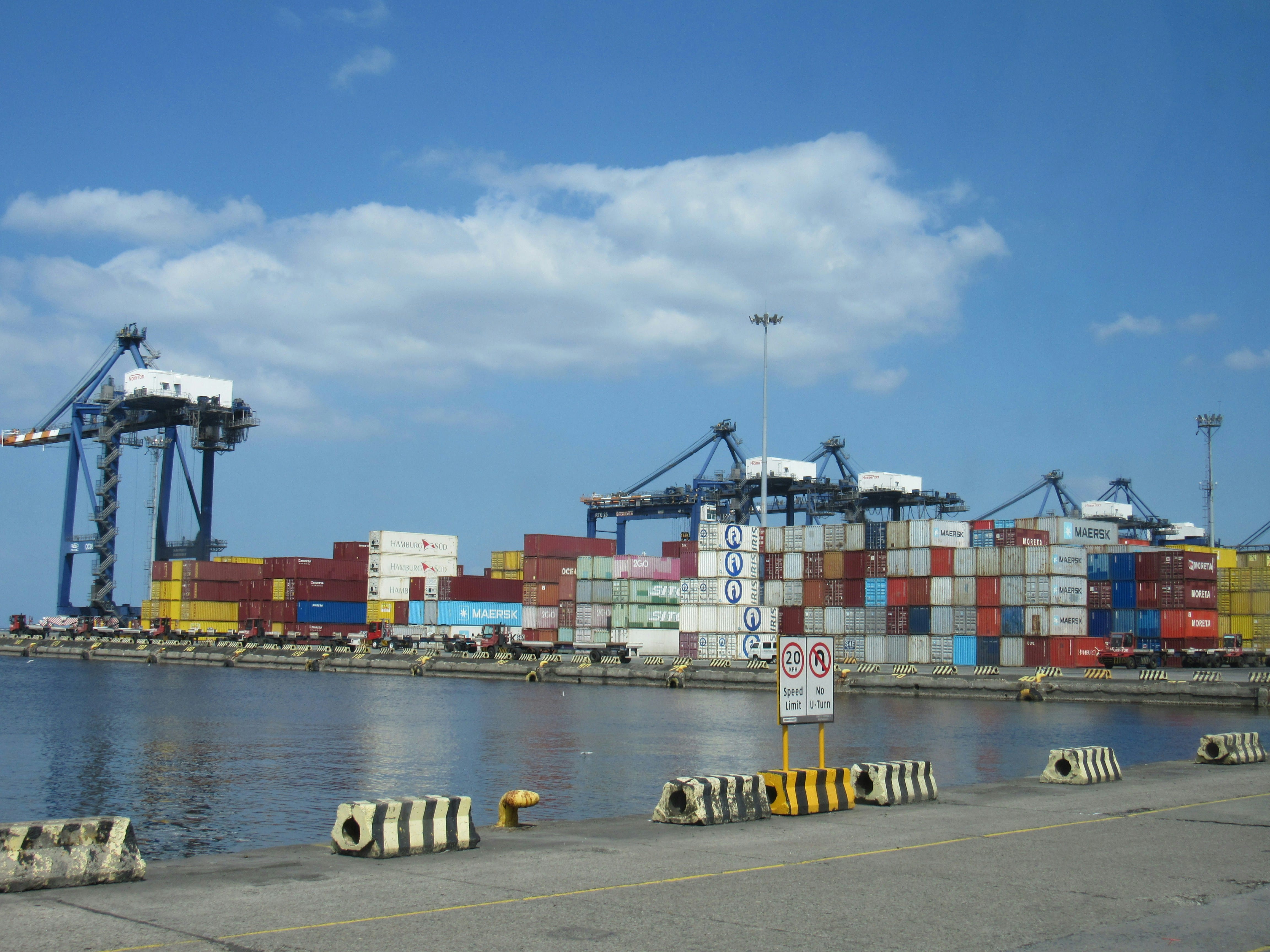 A modern shipping port with stacked multicolored shipping containers, large cargo cranes, and a fully loaded container ship docked at the facility under a clear blue sky with white clouds.