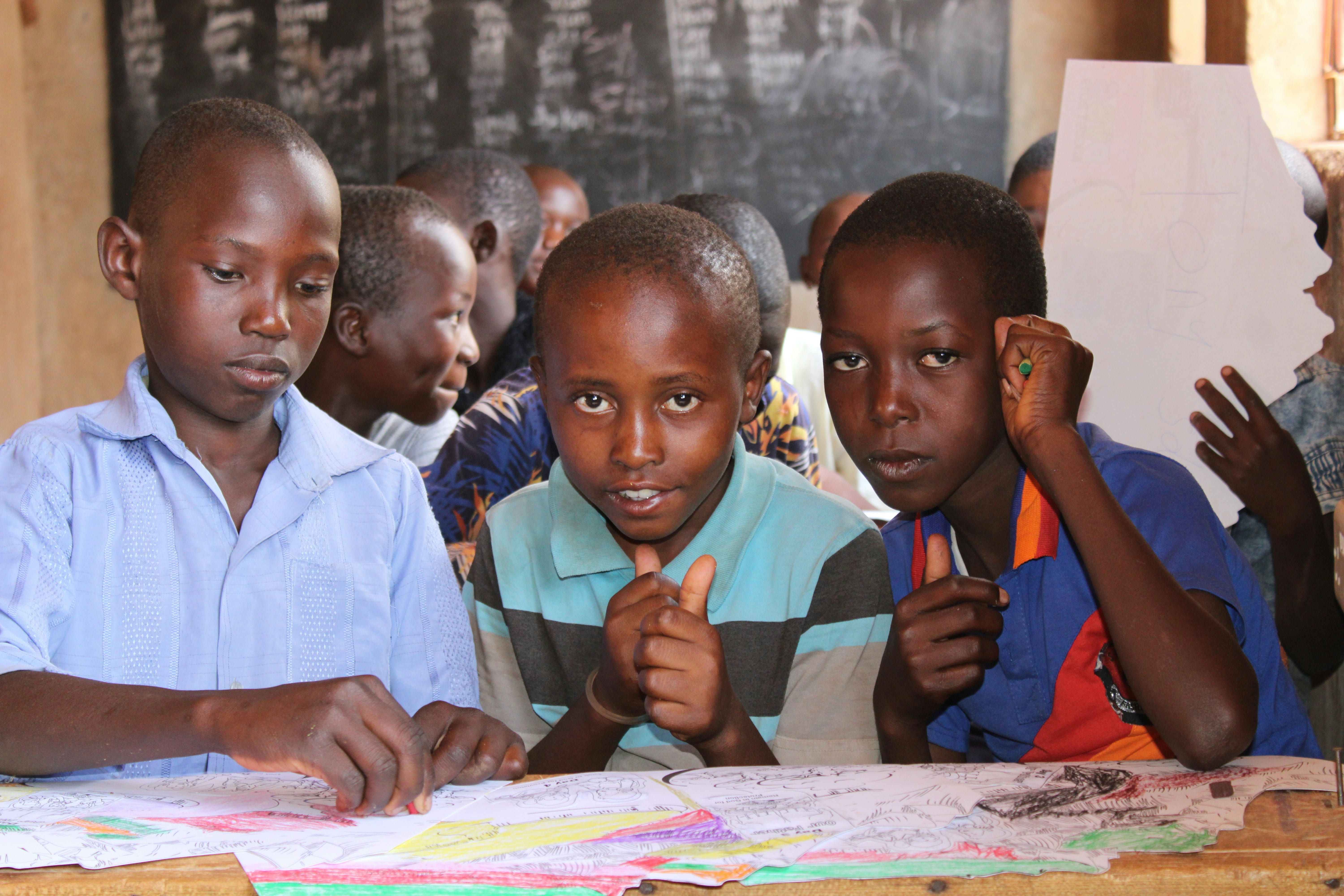 Three African adolescents sit at a wooden desk in a classroom, looking toward the camera with engaged expressions while studying from open books and papers, with additional students visible in the background.
