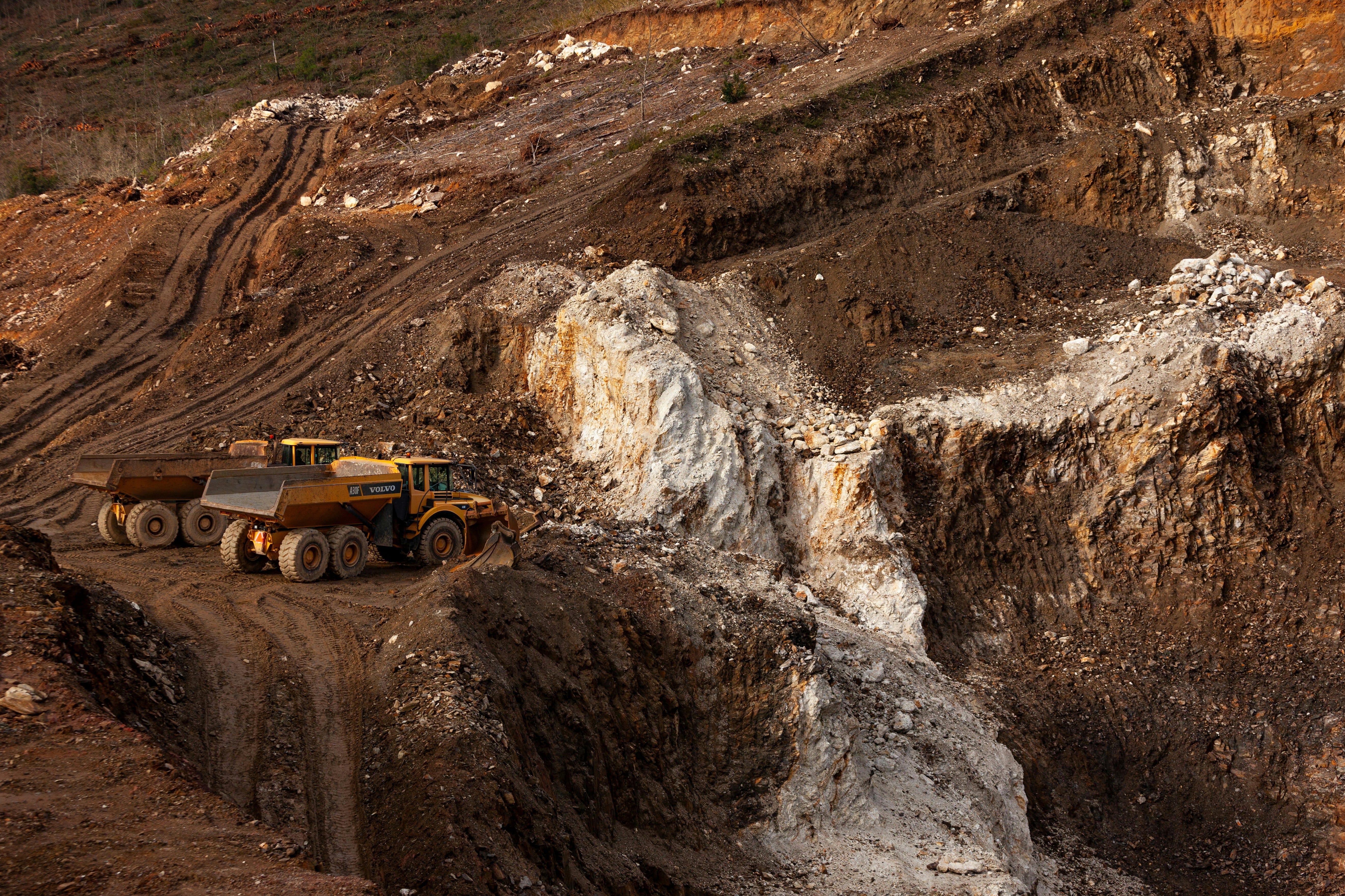 Aerial view of an active open pit mining operation showing an orange/yellow heavy-duty mining truck on a dirt road with exposed white mineral deposits and brown earth layers visible across the excavated landscape.