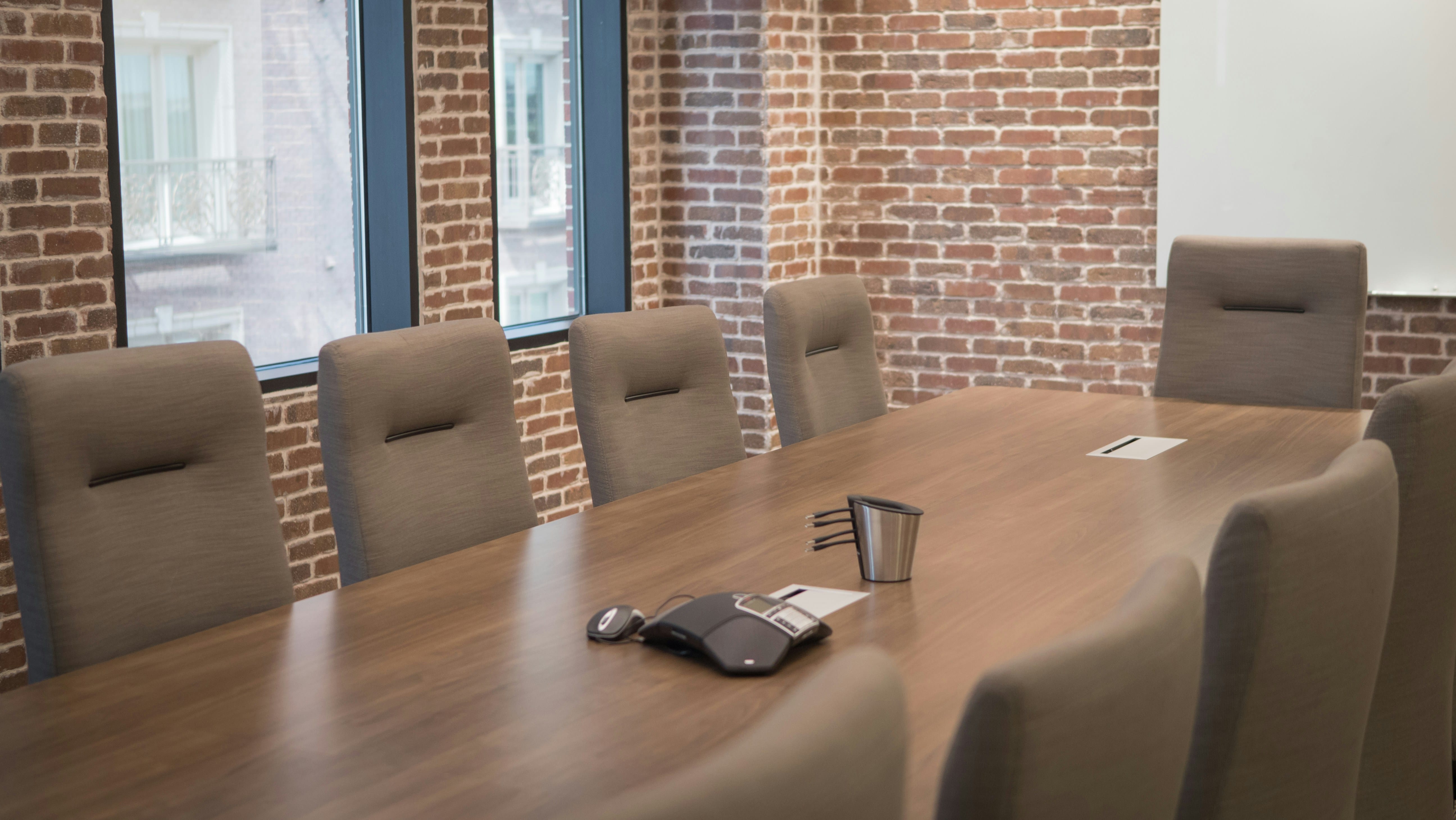 A wooden conference table in a brick-walled meeting room with six gray upholstered chairs positioned around it, a metal water pitcher and glass visible on the table surface, and windows with dark frames in the background.