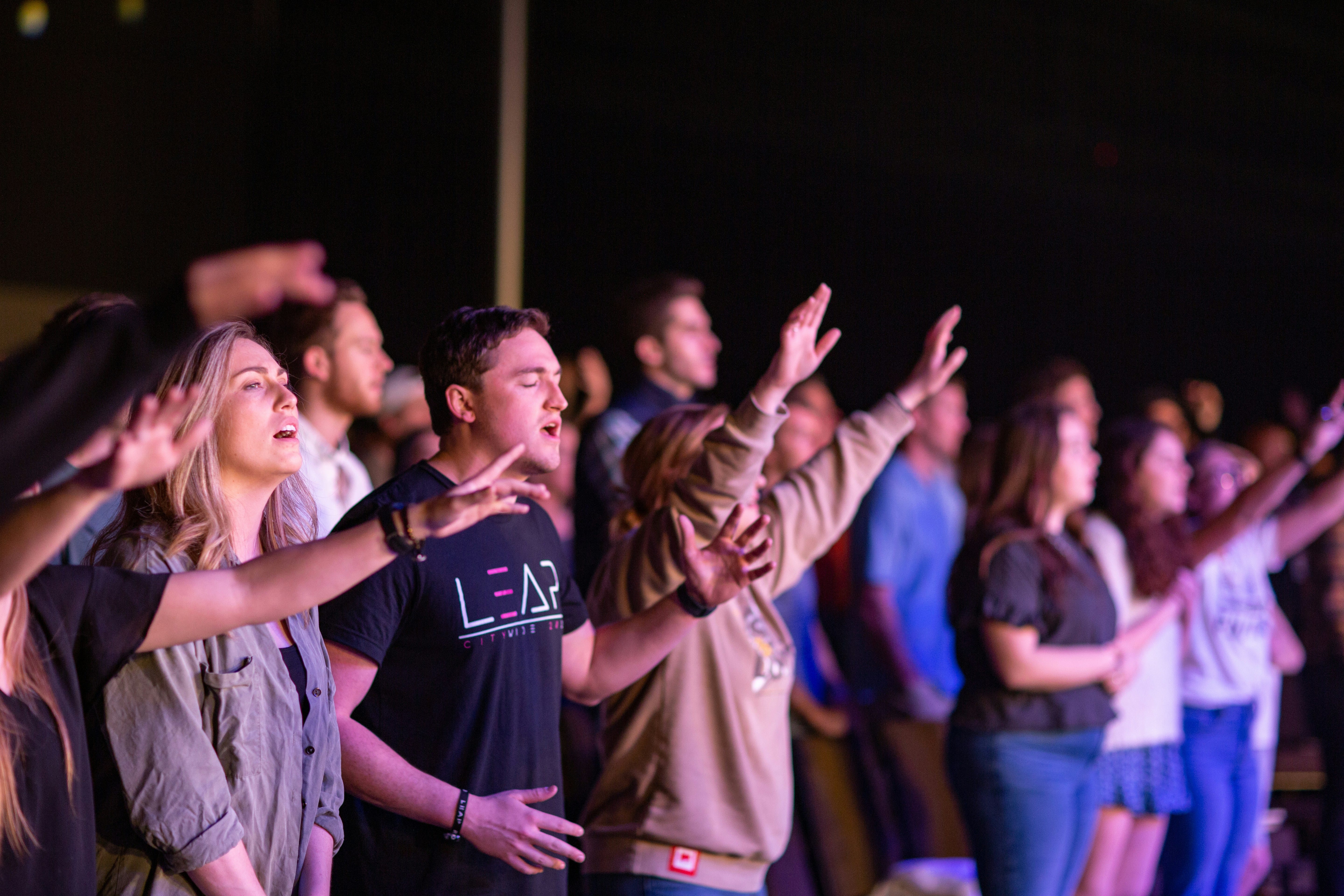 A group of young people with arms raised in worship during a contemporary religious gathering in an indoor venue, lit with purple and pink stage lighting, showing engaged participation in a community worship setting.