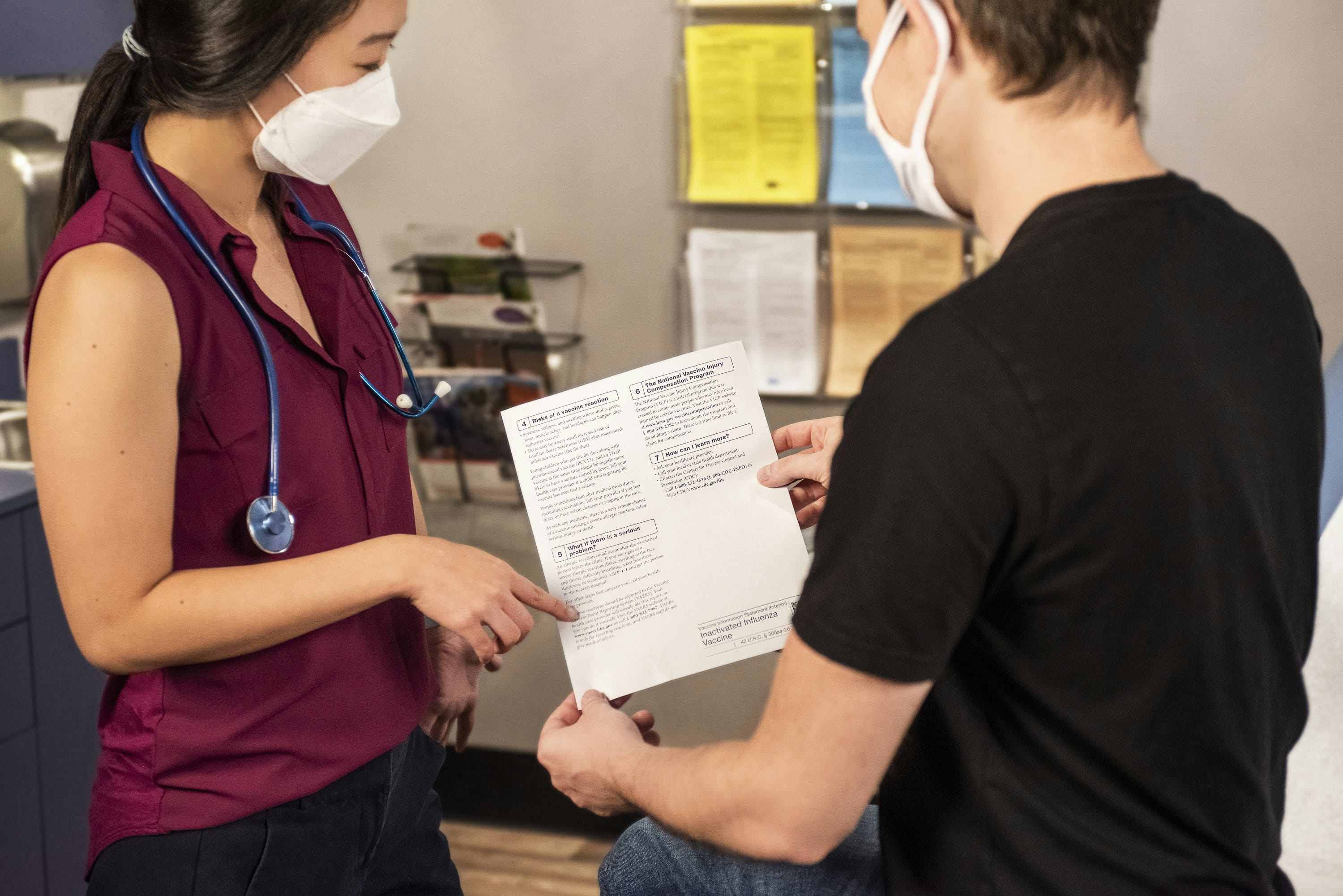 A healthcare professional wearing a maroon scrubs and stethoscope, standing and reviewing a document with another person in a medical office setting with visible shelving and wall-mounted materials in the background.