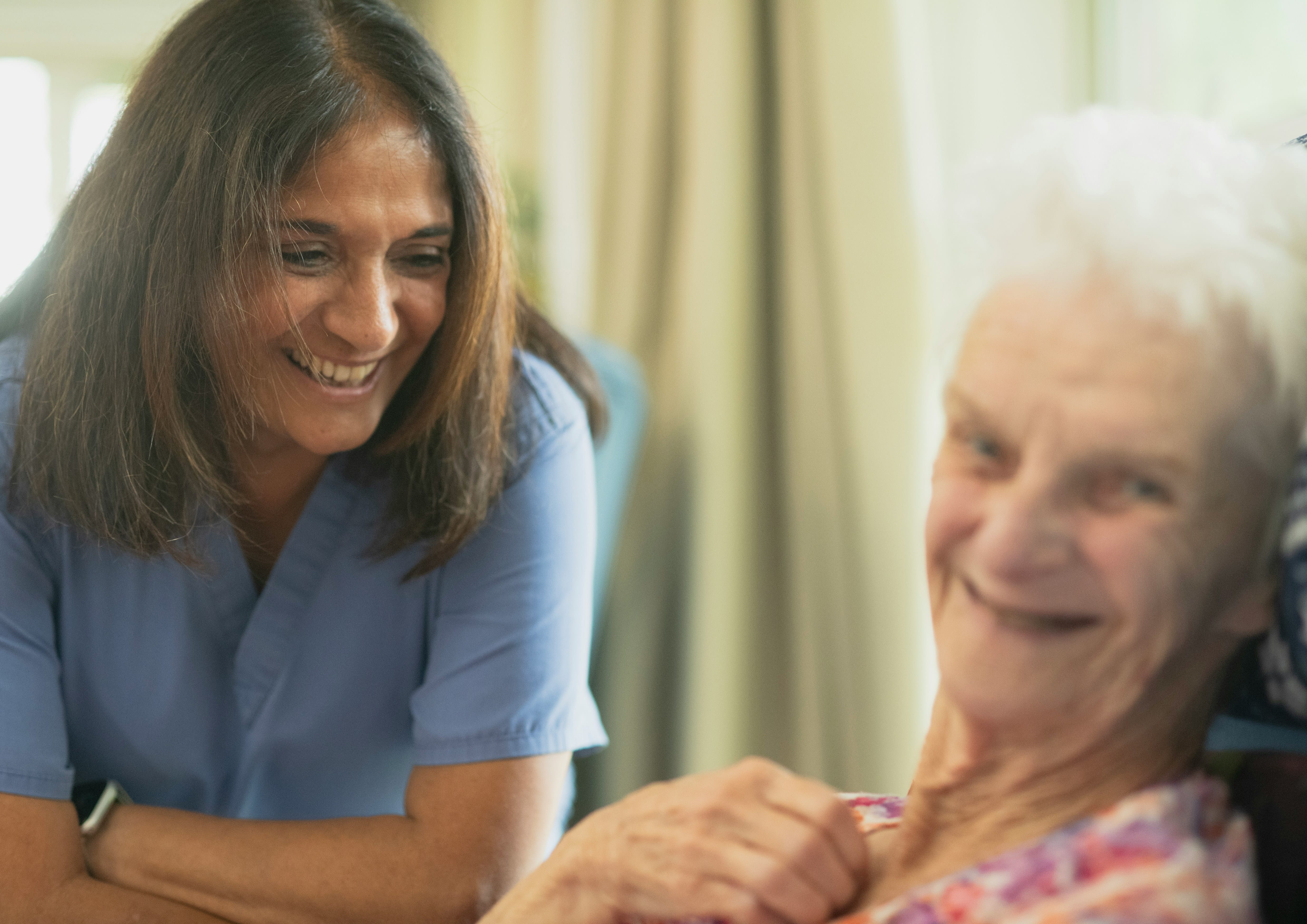 A healthcare professional in blue scrubs smiles while consulting with an elderly woman in a comfortable indoor setting, demonstrating a caring and supportive interaction.