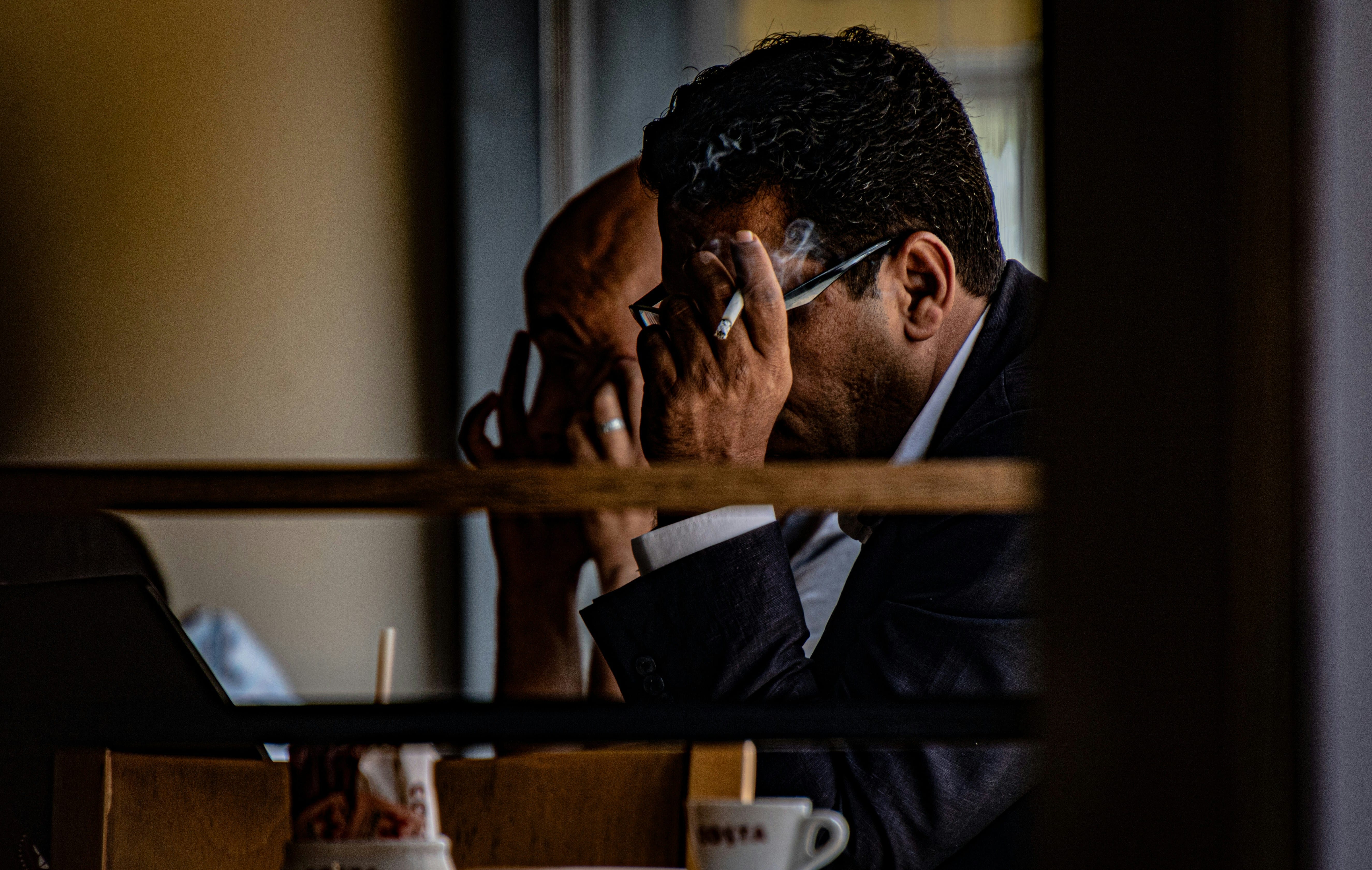 A professional man in a dark business suit sits at an office desk with his hand to his face in a contemplative pose, suggesting stress or concentration, with blurred office interior visible in the background.