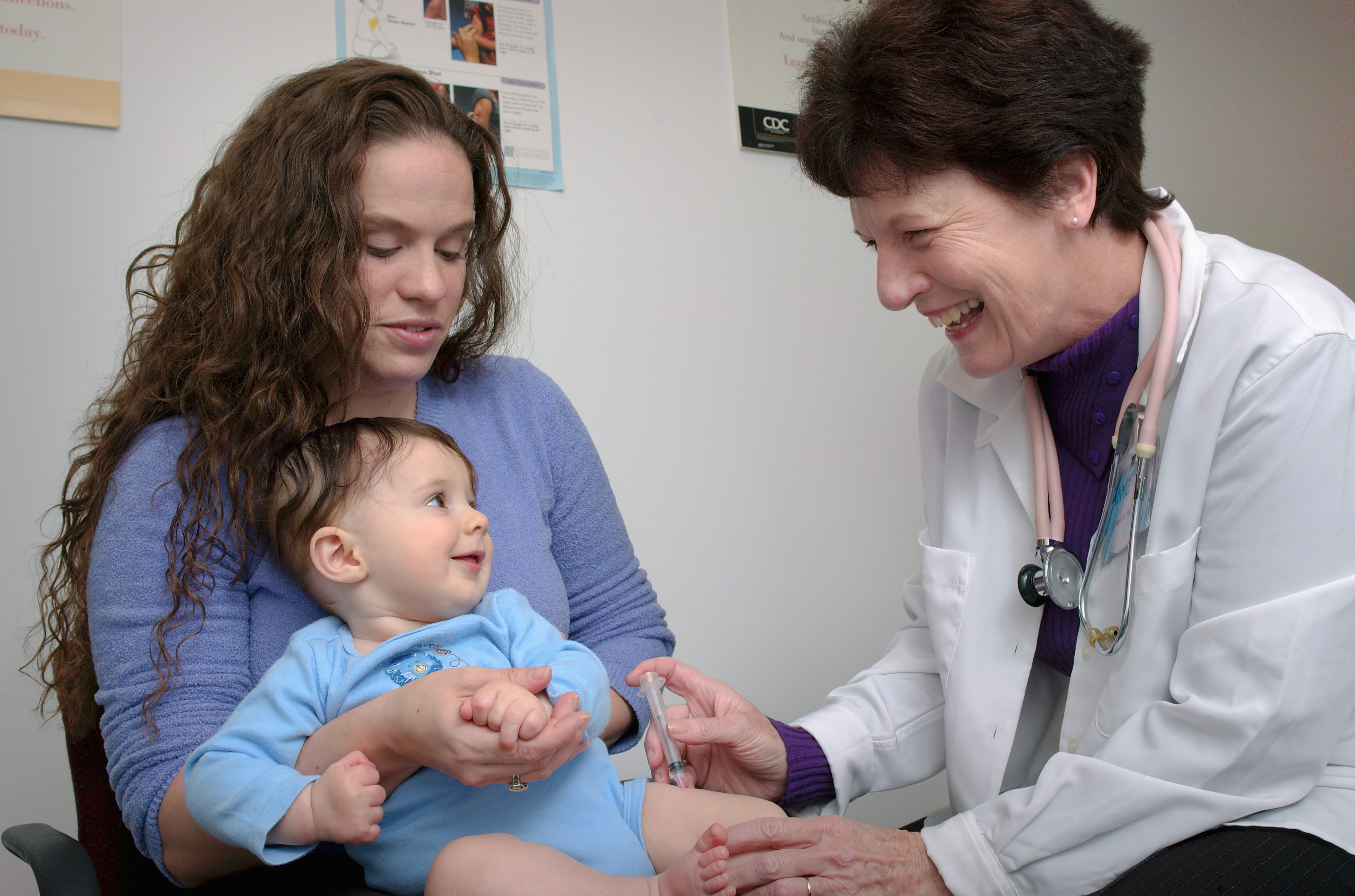 A pediatric doctor in a white coat examines a young child held by an adult woman in a clinical office setting, with the doctor smiling warmly at the patient during a consultation.