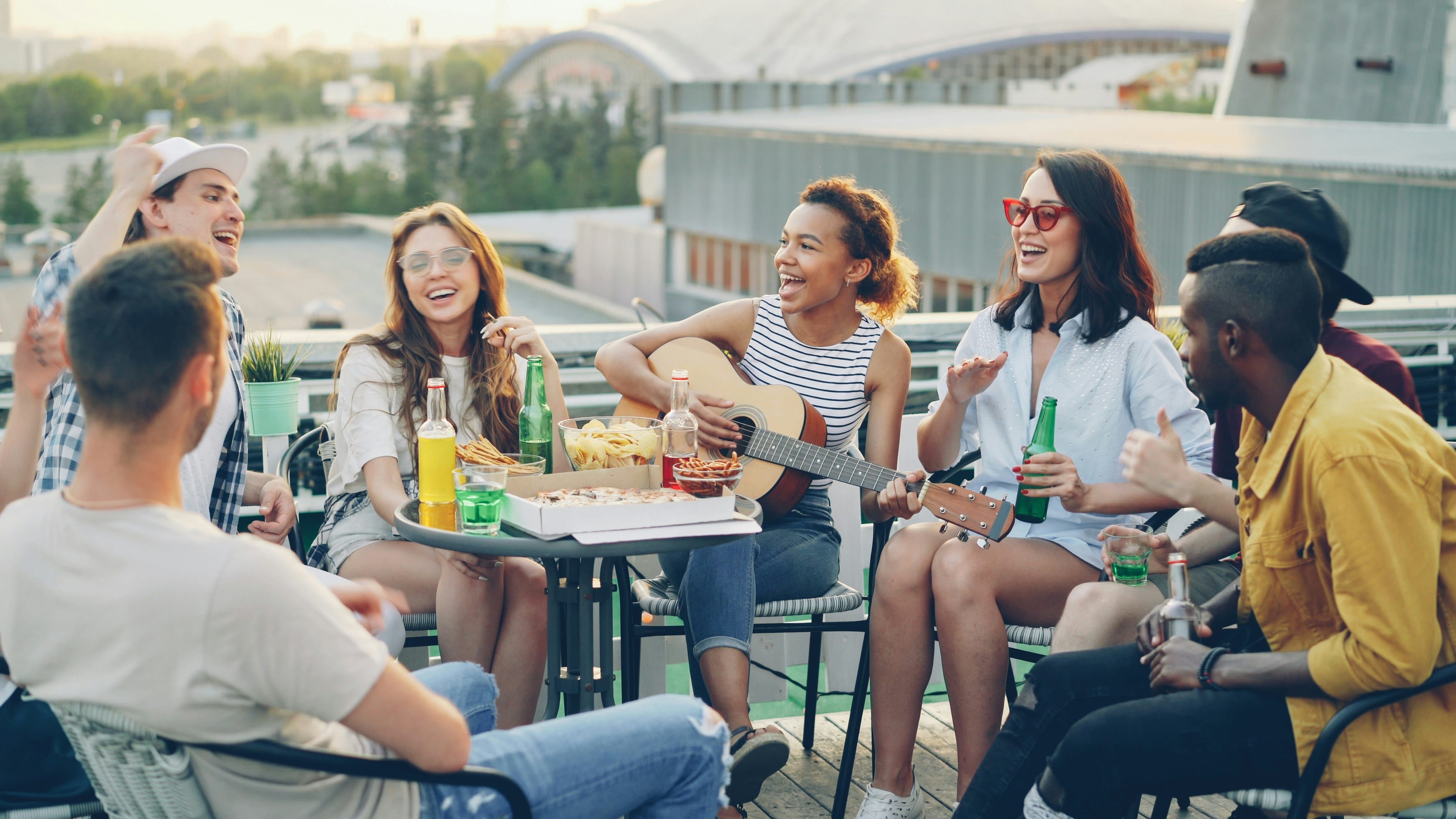 A diverse group of six people sitting in a circle outdoors on a rooftop or elevated urban space, engaged in conversation around a table with food and beverages, with a bridge and buildings visible in the background.