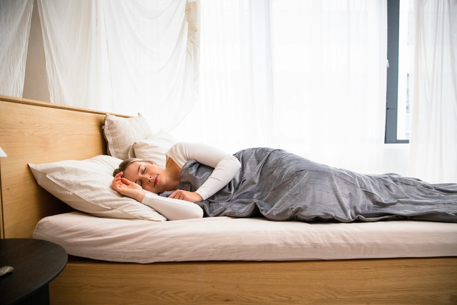 A person sleeping peacefully on a wooden bed with white linens in a bright, minimalist bedroom with natural light streaming through windows.