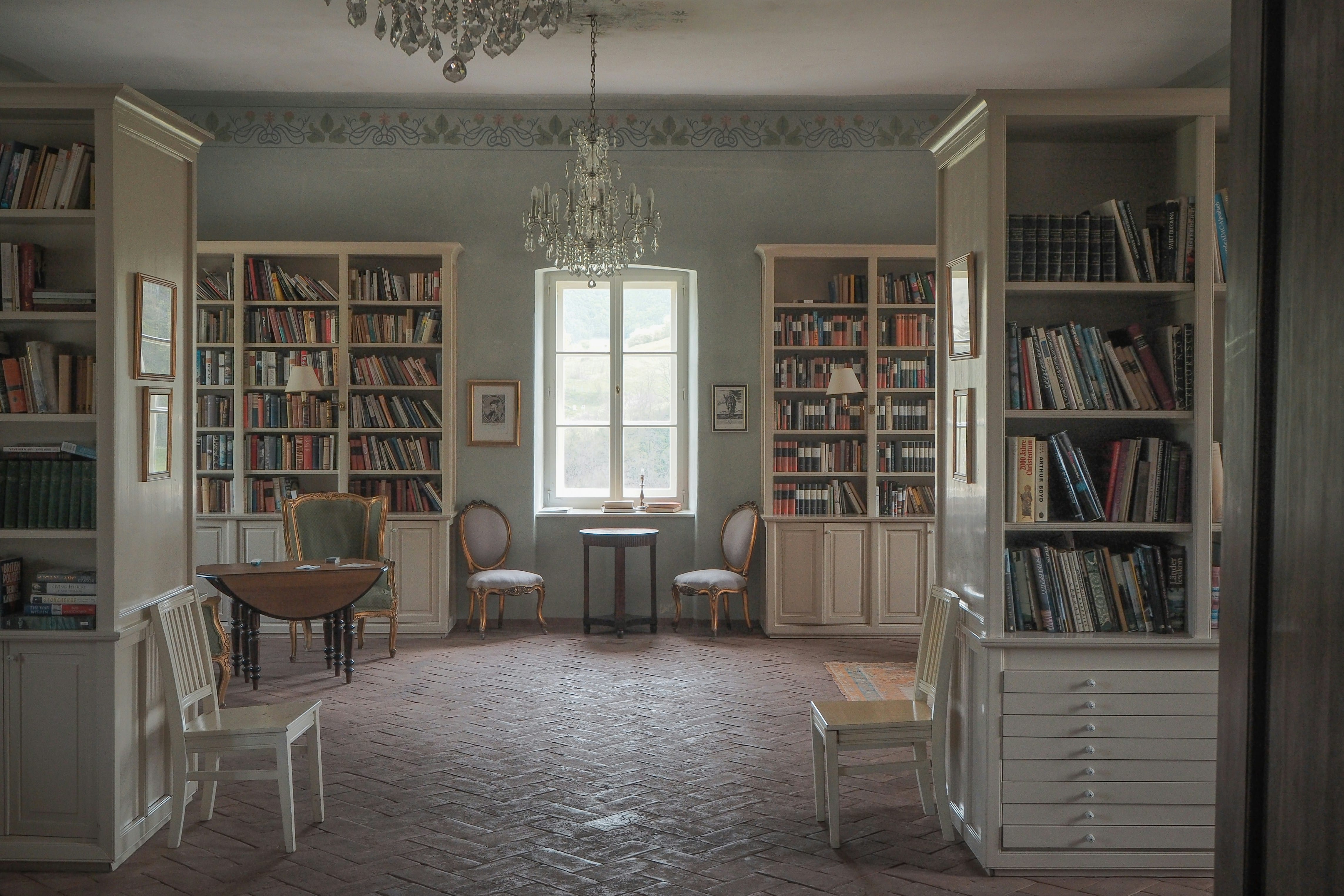 A traditional library room with floor-to-ceiling wooden bookshelves on multiple walls, a crystal chandelier hanging from the ceiling, two upholstered chairs positioned near a window with natural light, and patterned wallpaper, creating a scholarly and contemplative atmosphere.